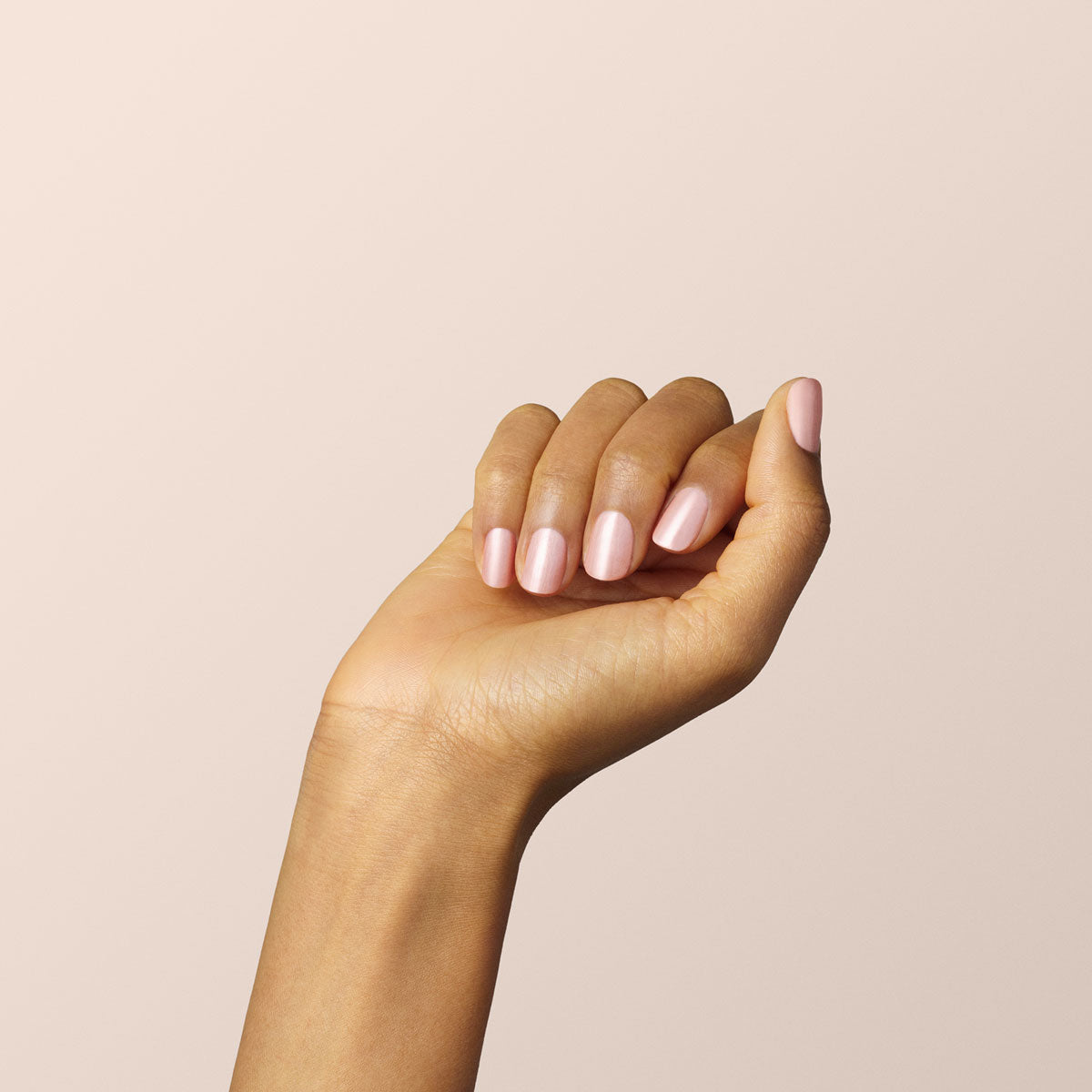 Une main aux ongles impeccablement manucurés vernis avec le gel Carnation de Manucurist, un rose nacré, présentée sur un fond beige clair uni, les doigts légèrement repliés pour révéler la carnation naturelle de la peau.