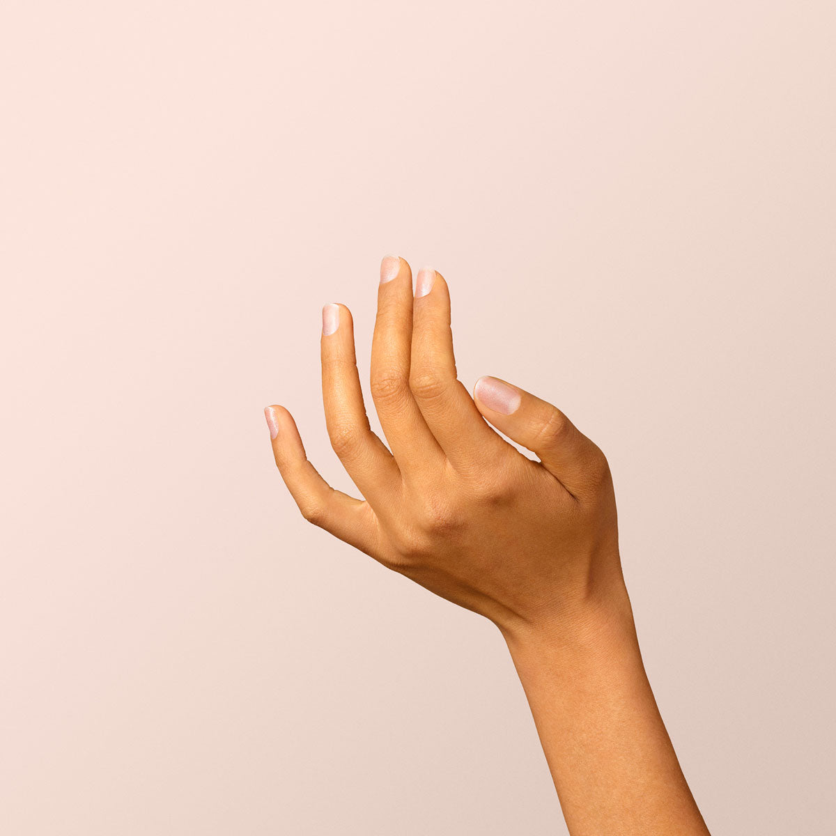 A hand with medium brown skin is elegantly raised before a light beige background, its well-groomed nails adorned with Diamant, a subtle silver polish by Manucurist, and no accessories in sight.
