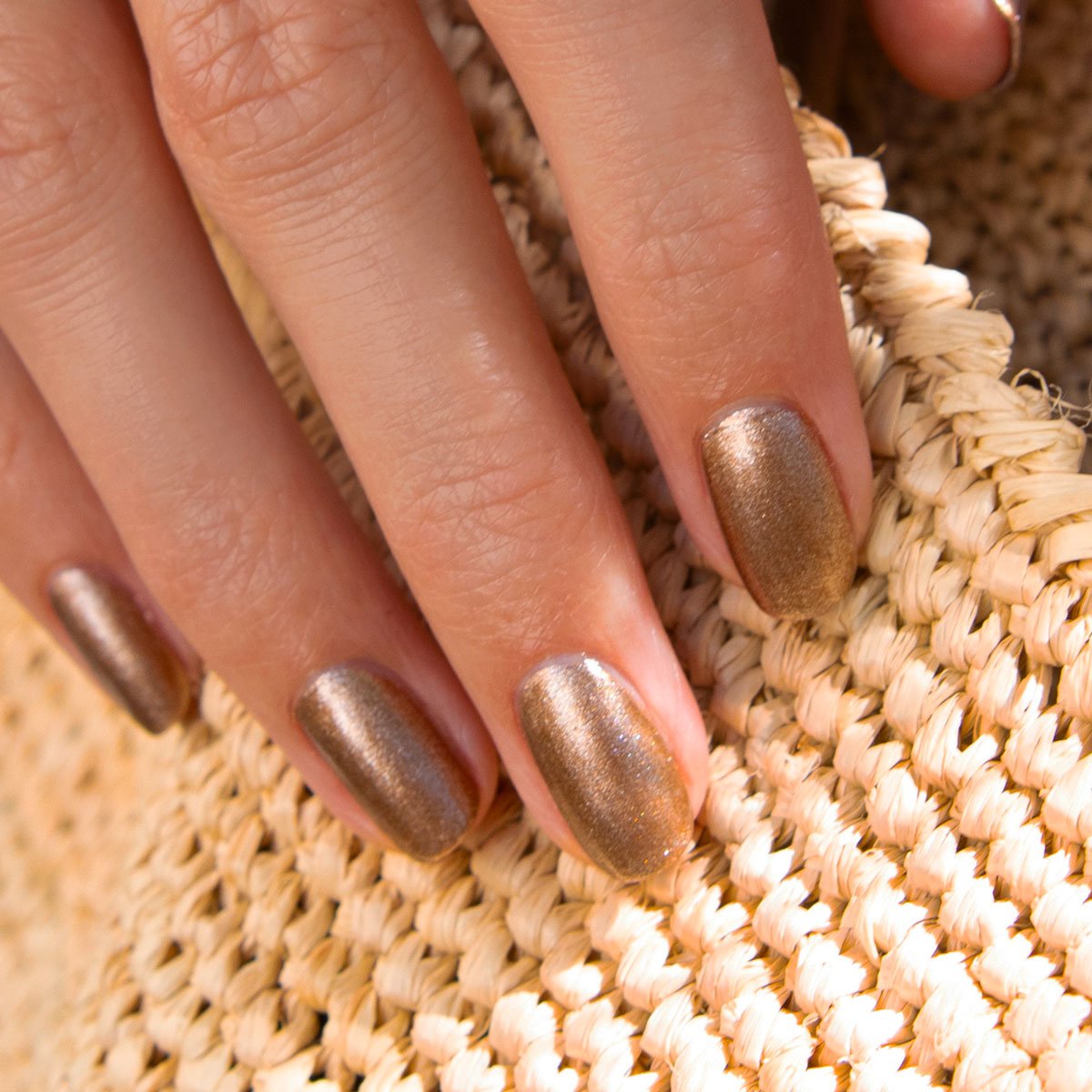 Gros plan sur une main aux ongles courts et impeccablement manucurés avec le vernis gel Bronzé de Manucurist, posée sur une surface en paille tressée.