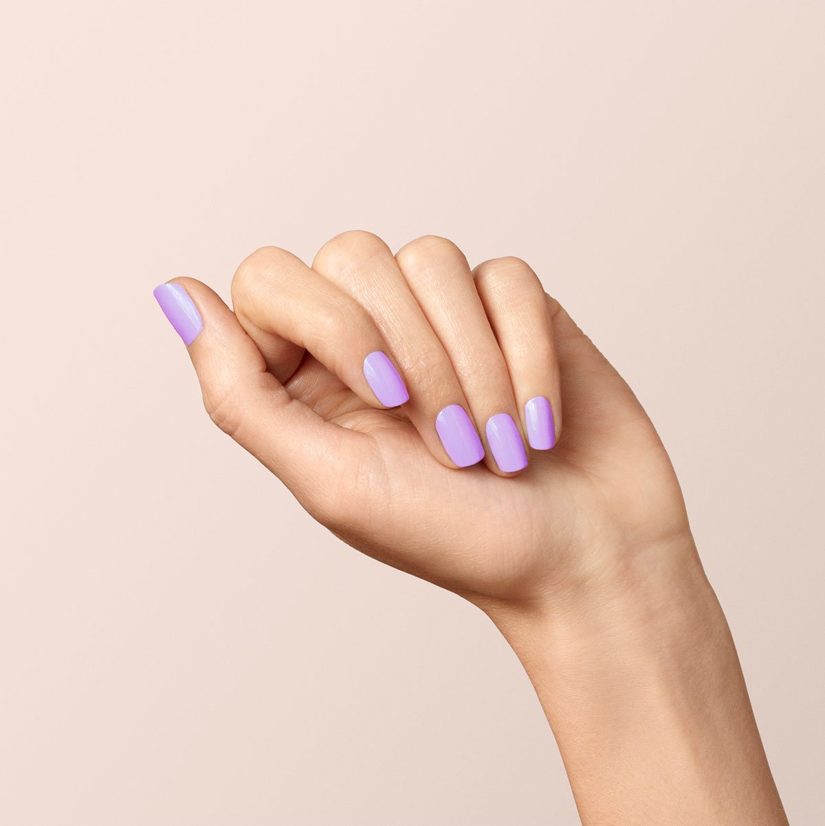 A hand with light skin is pictured against a beige background, nails painted in the glossy Lisa Lilas shade from Manucurist, fingers gently curved inward.