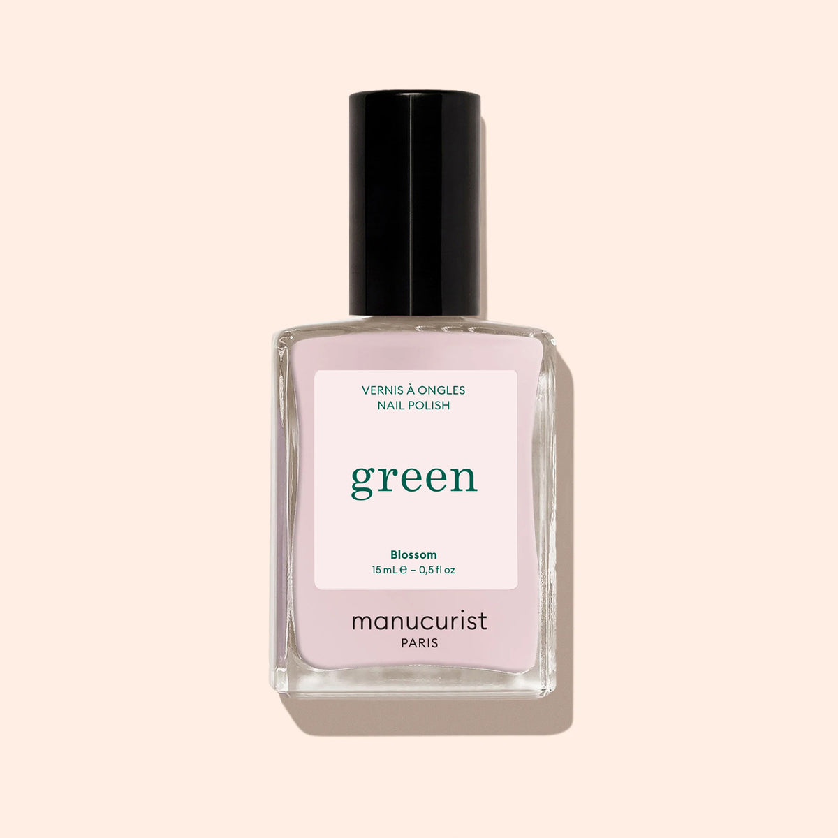 Un flacon de vernis à ongles Manucurist dans la teinte « Blossom », affichant une couleur rose clair crémeuse, un bouchon noir et un flacon carré en verre transparent de la gamme Green, est présenté sur un fond beige.