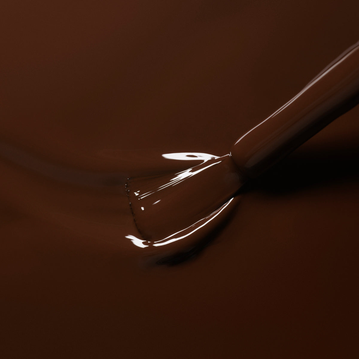 A close-up of a nail polish brush dipping into Manucurist’s Clove, a glossy chocolate brown gel polish, with smooth, shiny liquid coating the bristles.
