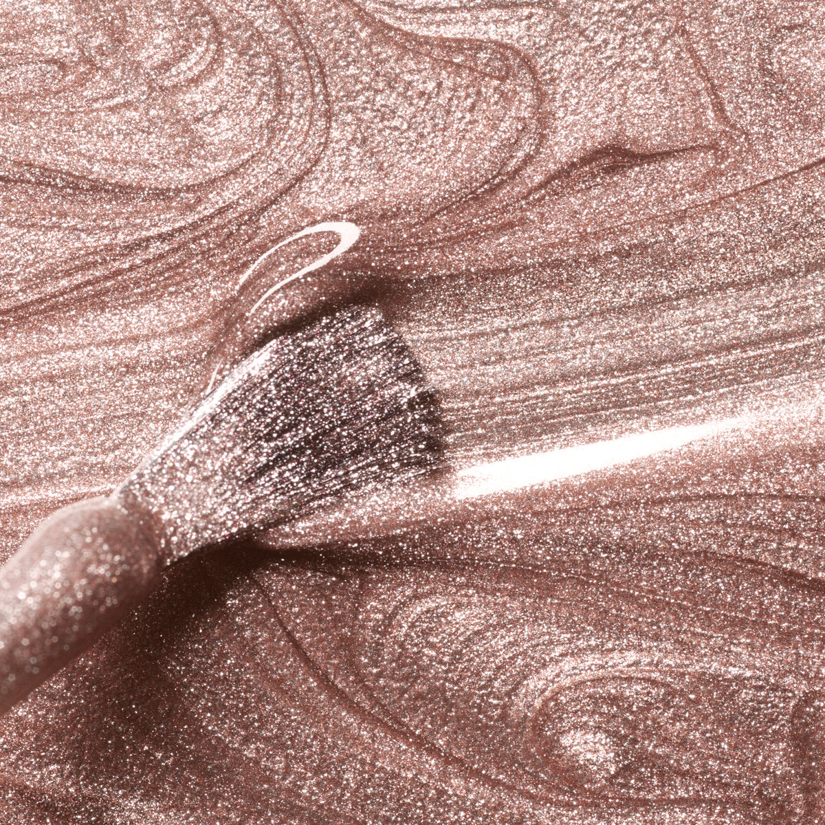 A close-up of a brush dipped in Manucurist Cosmic Rose gel polish, swirling shimmery champagne pink shades and creating textured patterns in the glittery liquid.