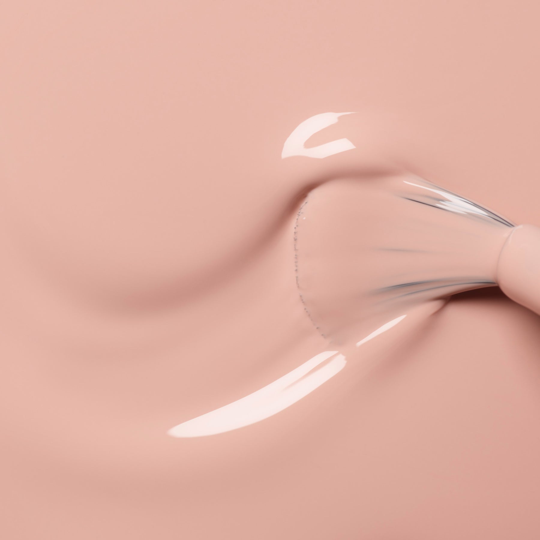A close-up of a brush swirling Manucurist Dune foundation, a creamy light pink shade with orange undertones, on a smooth surface, creating a glossy texture and softly blurred streaks.