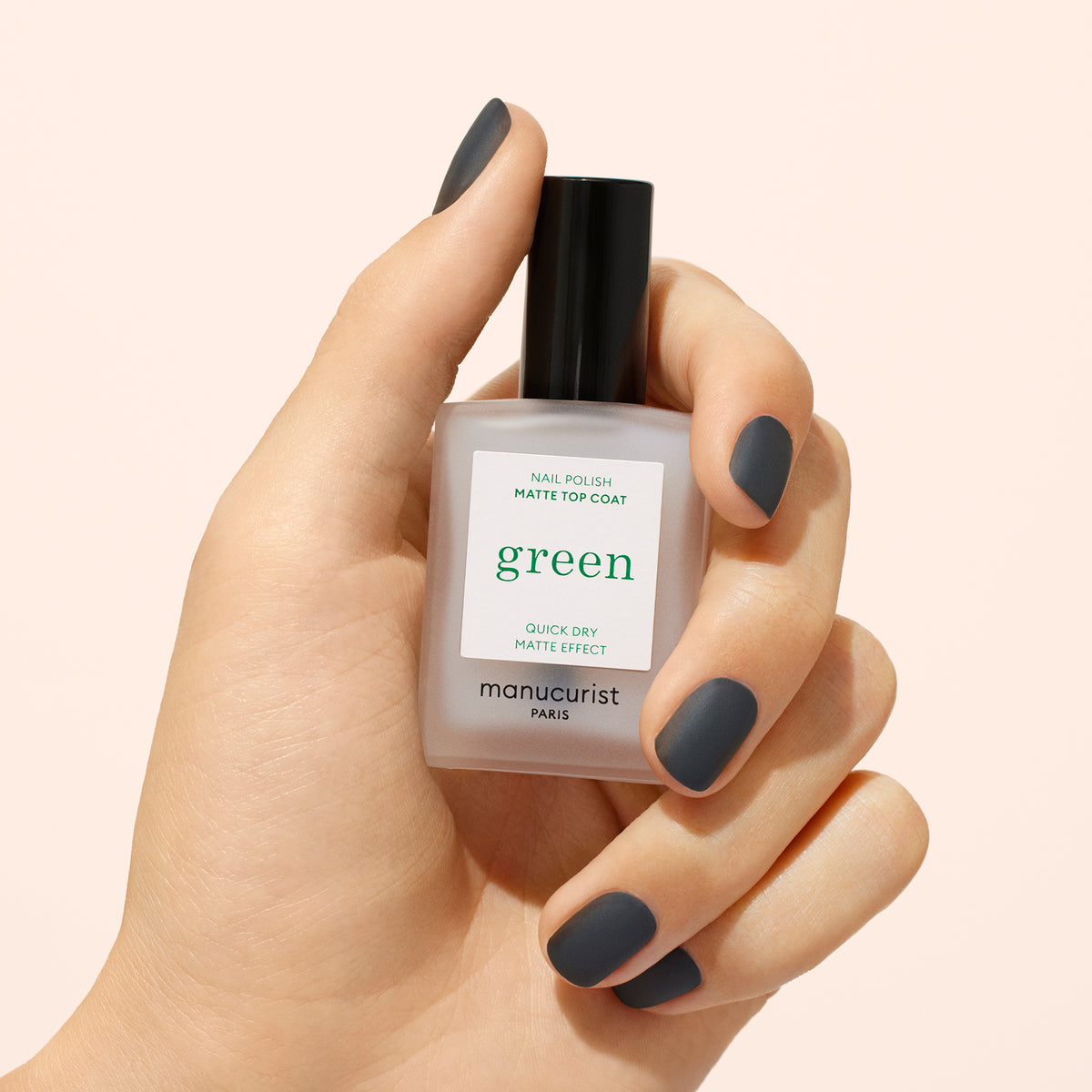 Une main aux ongles courts, mats et gris foncé tient un flacon de Matte Top Coat Manucurist, un vernis d&