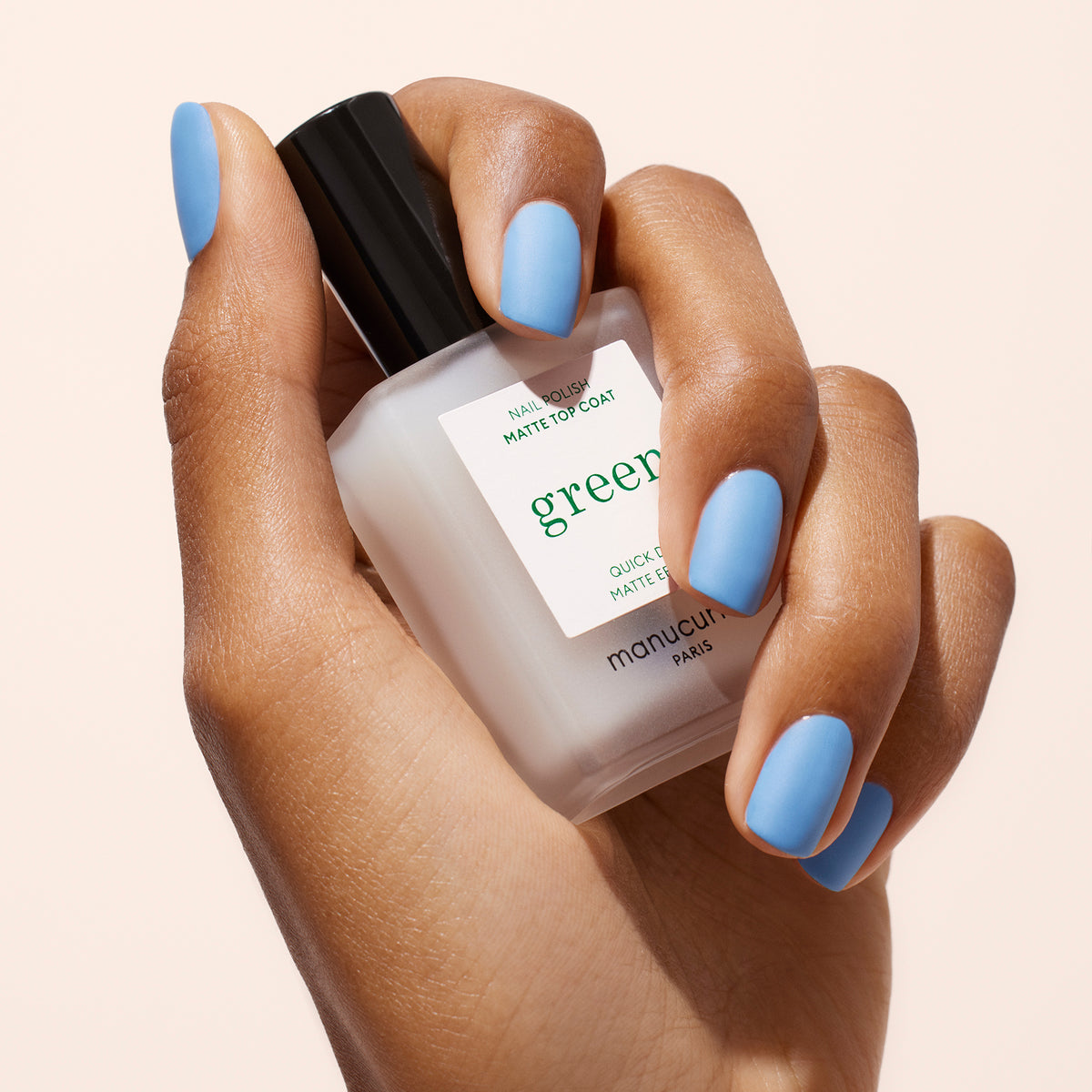 Une main aux ongles bleu clair tient un flacon givré de vernis à ongles Manucurist Matte Top Coat avec un bouchon noir, parfait pour une manucure écoresponsable, sur fond clair.