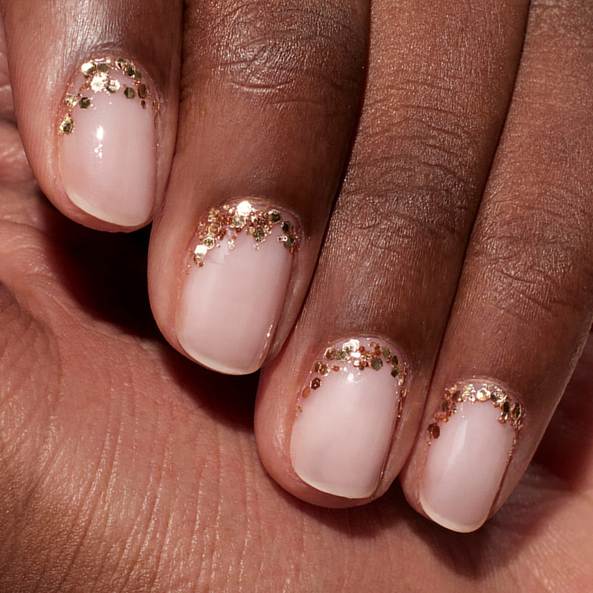 A close-up of a hand with natural pink nails displays elegant nail art, featuring Manucurist Rose Gold Biodegradable Glitter accents near the cuticles for a subtle, eco-friendly look.
