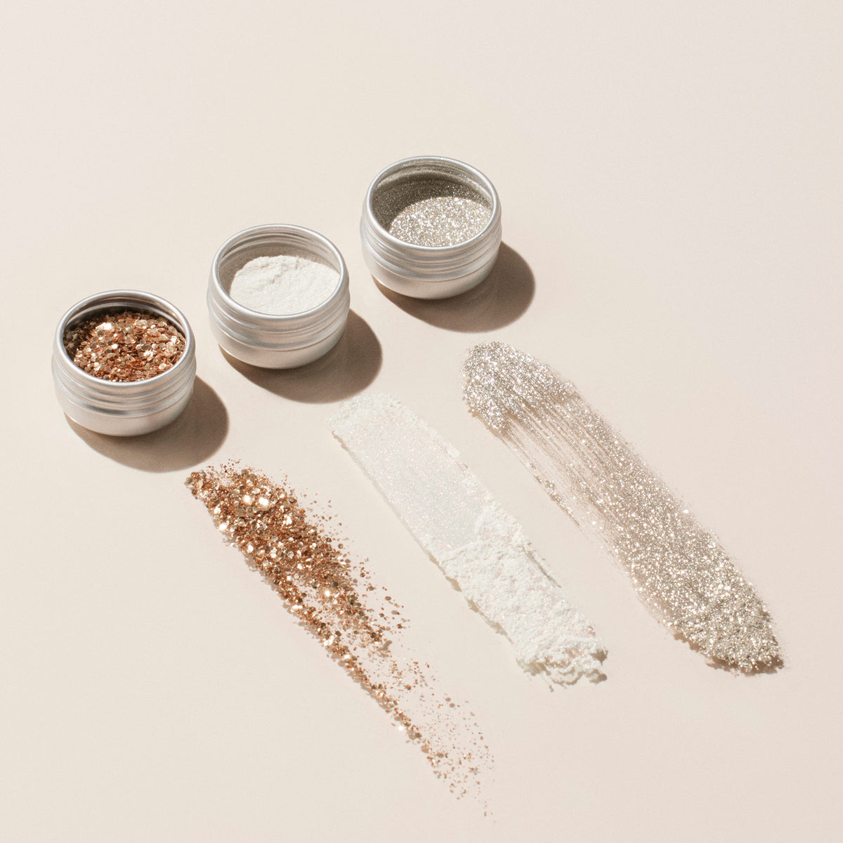 Trois petites boîtes métalliques du Glitter Trio de Manucurist — Rose Gold, Glazed et Mirror — sont alignées, avec chaque poudre swatchée sous sa boîte. Parfait pour ajouter de l’éclat aux créations de vernis à ongles.