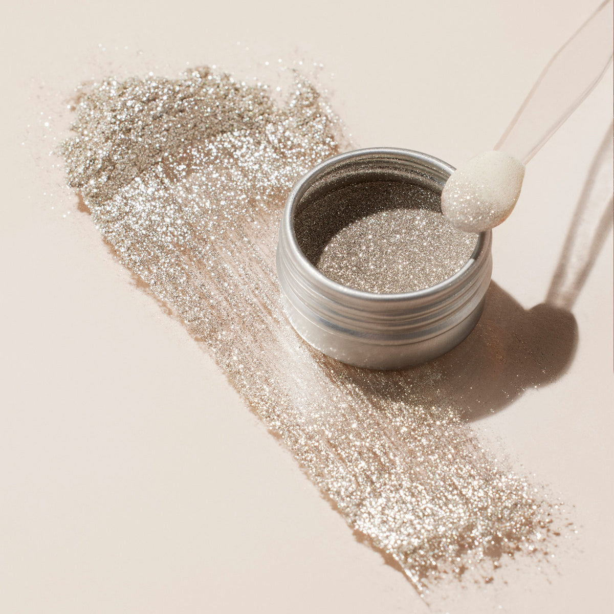 Un pot ouvert de Manucurist Glitter Trio—Rose Gold, Glazed, Mirror—est posé sur une surface claire, avec un peu de poudre renversée. Un applicateur à embout mousse repose sur le bord, prêt à ajouter des poudres à effet scintillant à votre look.