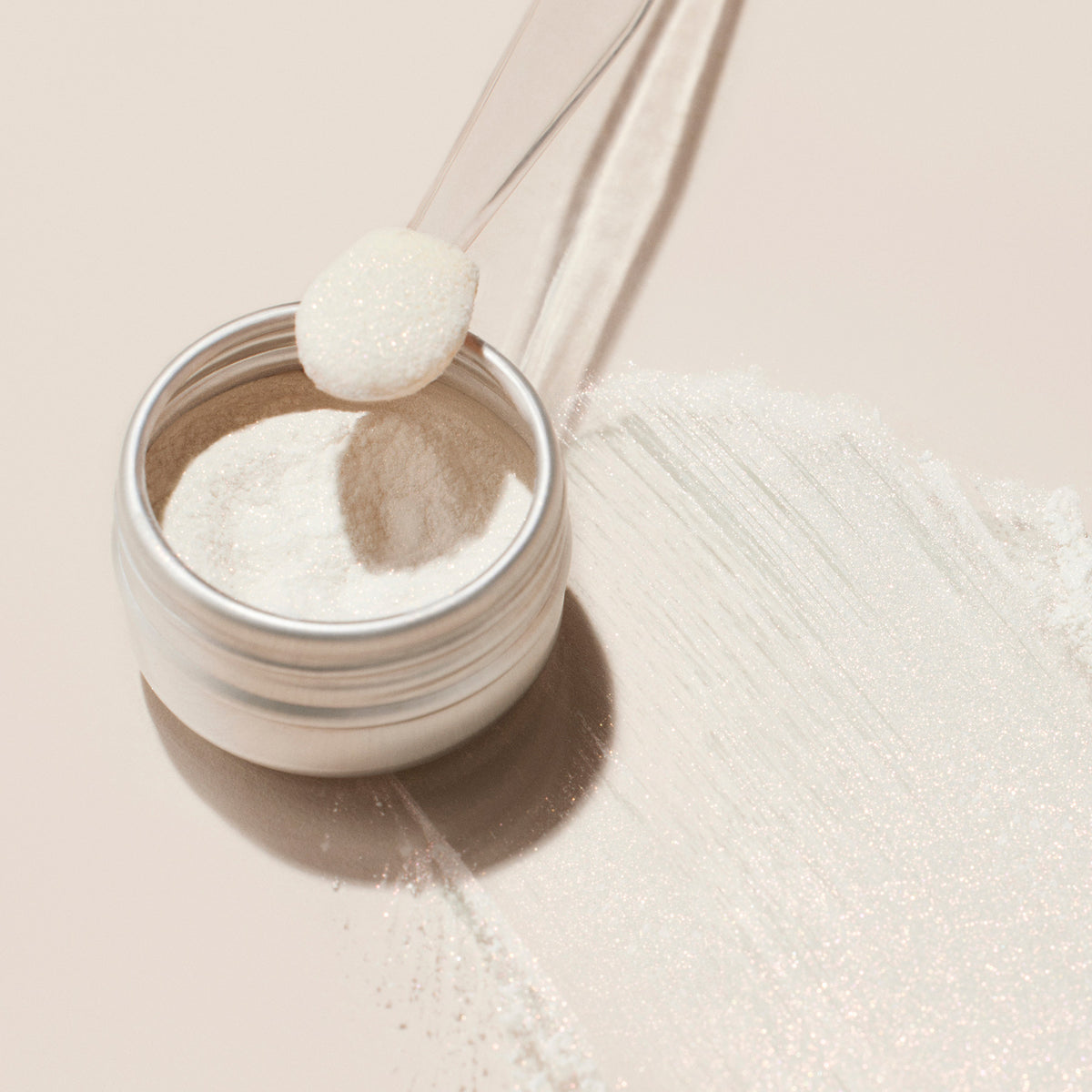 Un petit récipient en métal du Glitter Trio Manucurist (Rose Gold, Glazed, Mirror) est posé sur une surface beige. Une cuillère en plastique transparente contient de la poudre fine, avec une traînée de poudre à effet pailleté présentée à côté du récipient.