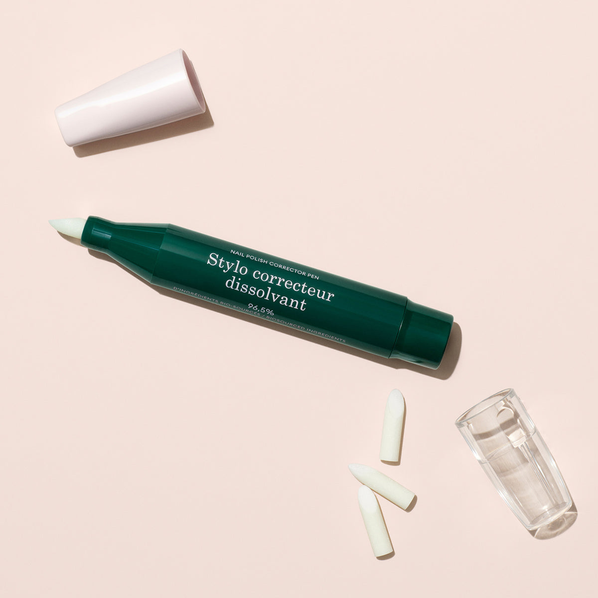 Le Manucurist-test + Stylo Correcteur pour vernis gel, présenté en vert sans son capuchon blanc, comprend deux embouts de rechange et un petit capuchon transparent, le tout disposé sur une surface beige clair.