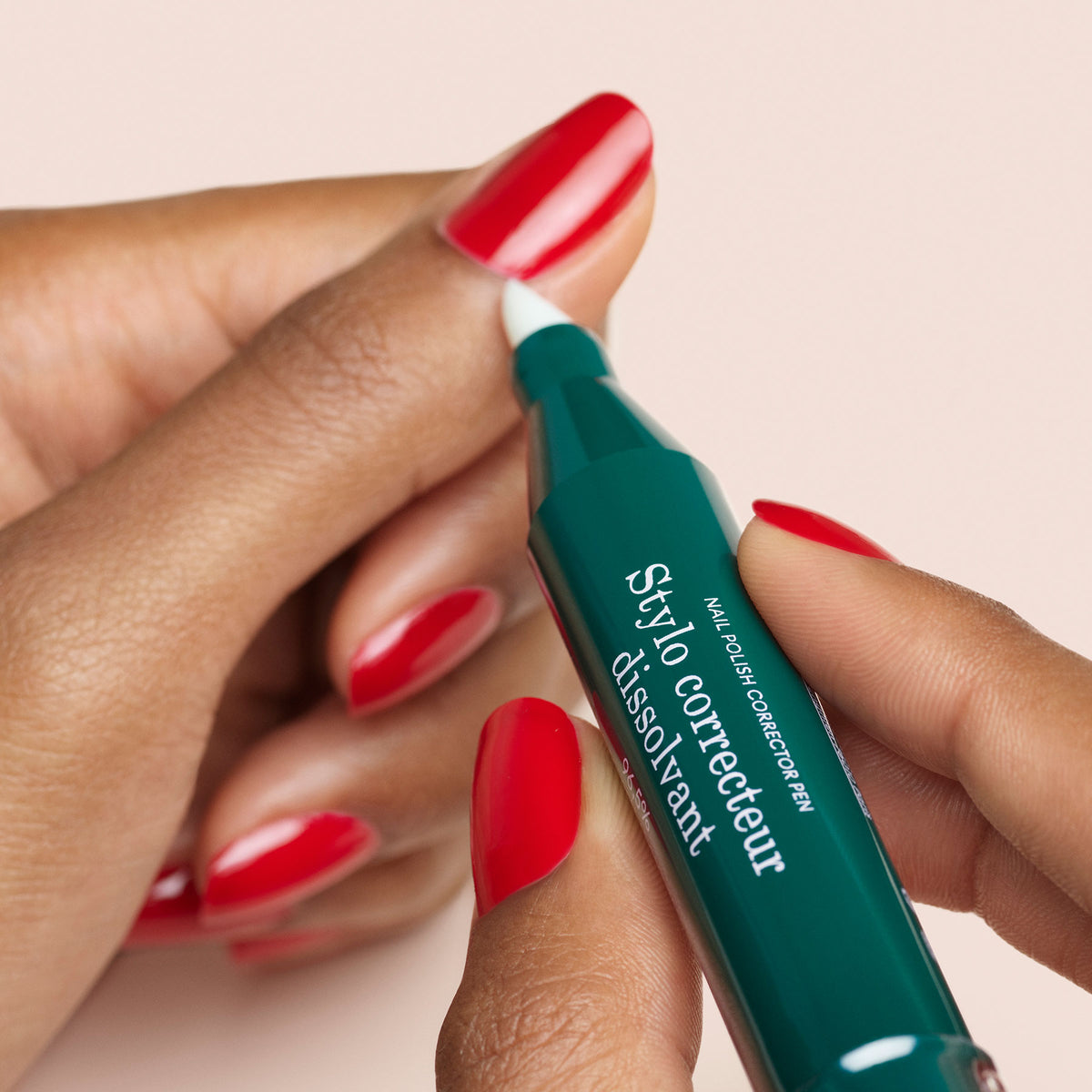 Une personne utilise le stylo correcteur Manucurist-test + pour nettoyer le vernis gel rouge sur ses ongles, sur un fond rose clair.