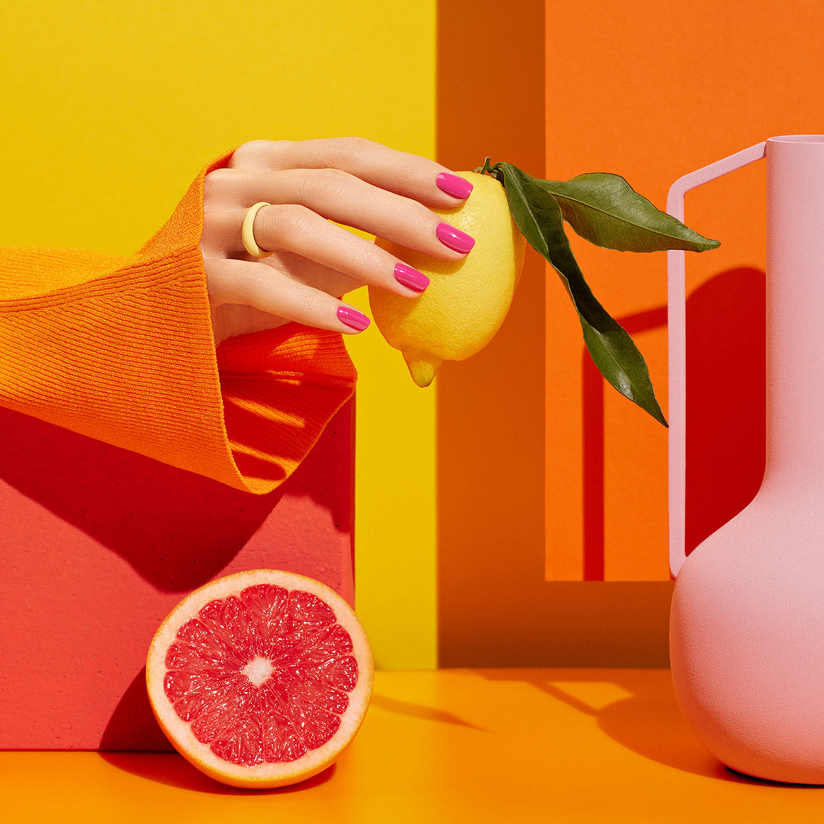 A hand with Manucurist’s Pétula bright pink polish and a gold ring holds a lemon against a vivid yellow-orange background, joined by a grapefruit half, pink vase, hints of green, and an orange sleeve for a lively scene.
