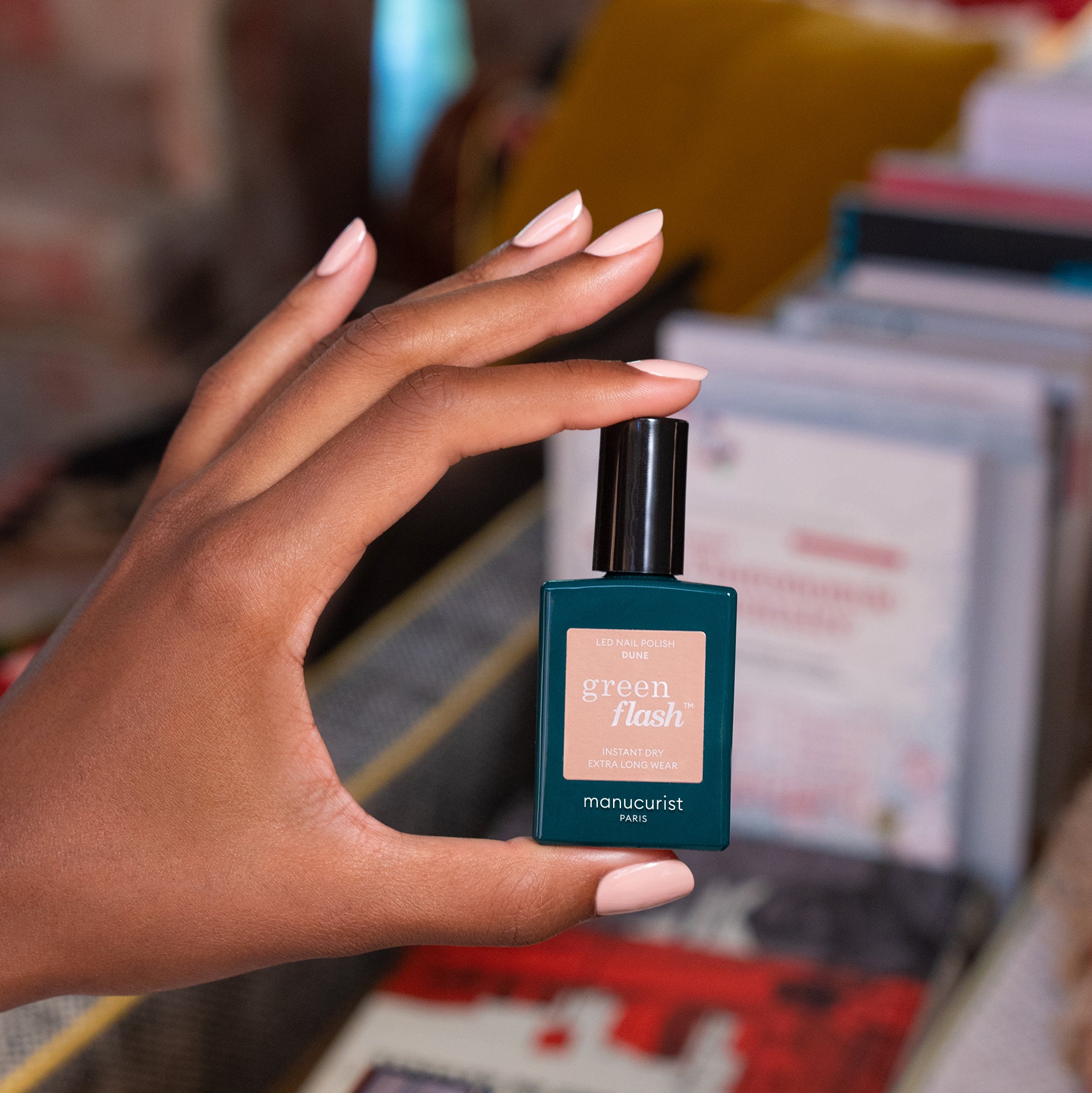 A hand with neatly manicured nails holds a bottle of Manucurist Dune polish in sandy beige, posed against a blurred backdrop with books and papers.