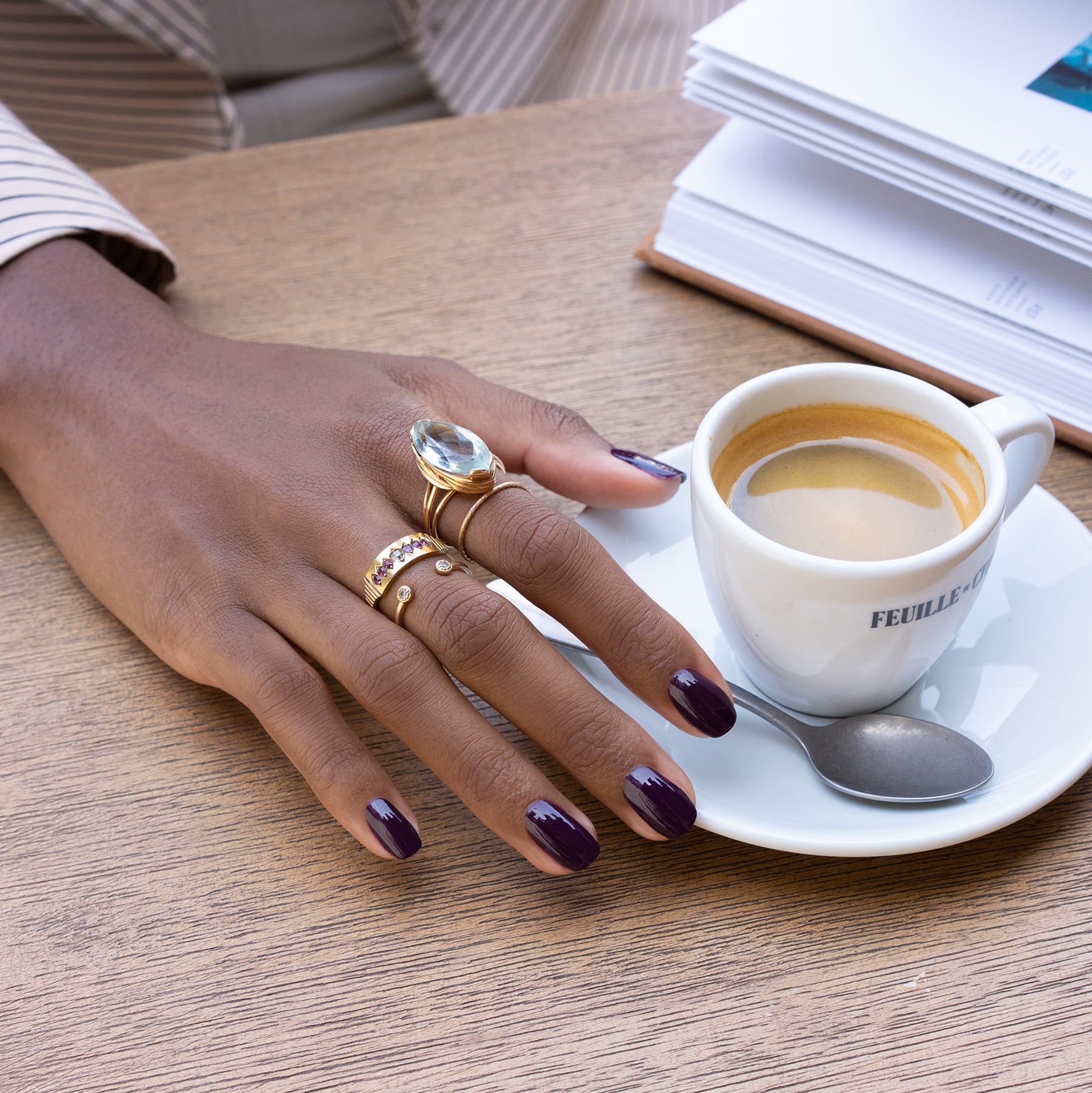 A hand with Manucurist’s Aubergine gel polish and multiple gold rings rests on a wooden table next to an espresso, a spoon, and an open notebook.