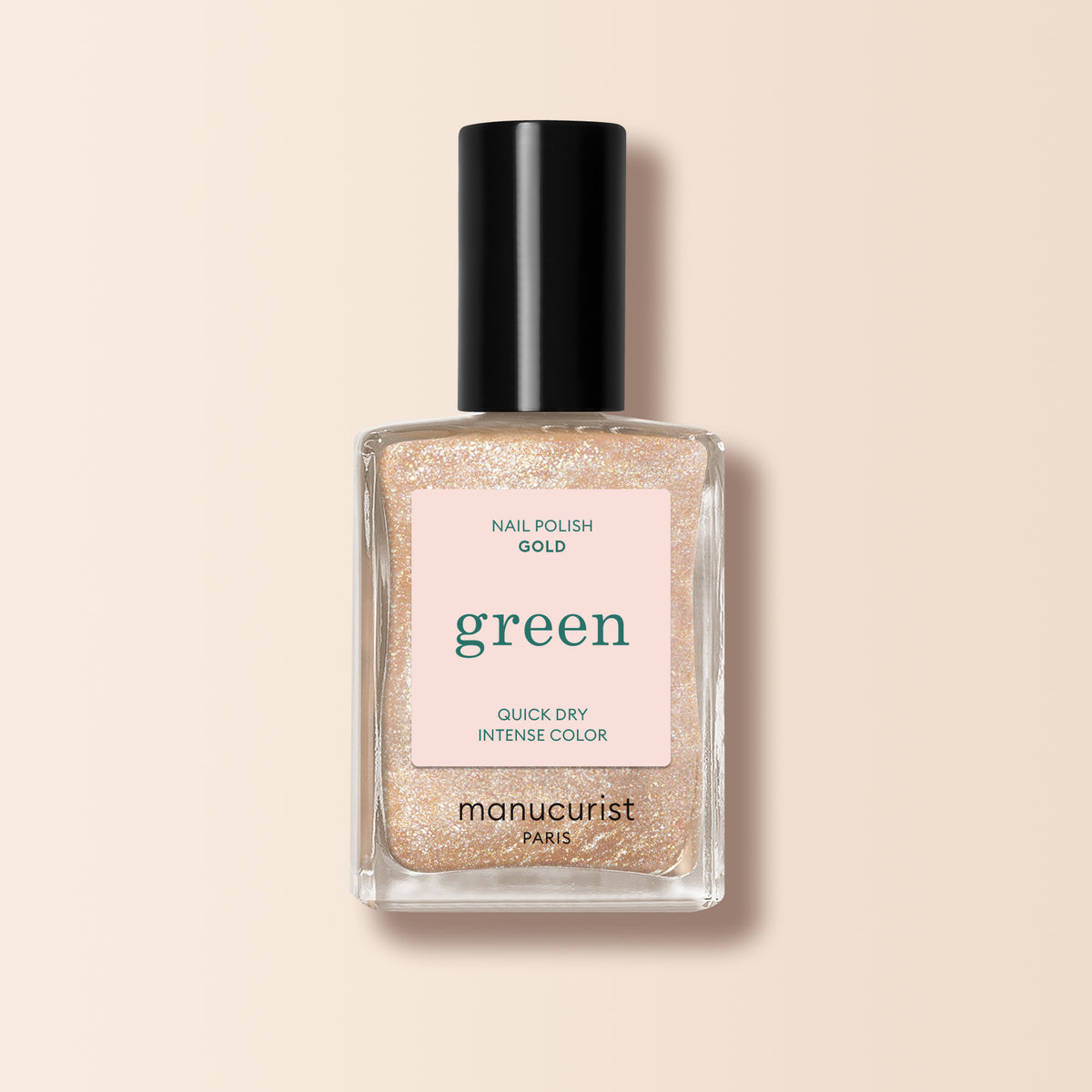 A bottle of Manucurist Gold nail polish from the Green range, with a black cap and a pink label stating "green," "nail polish gold," and "quick dry intense color," displayed on a beige background.