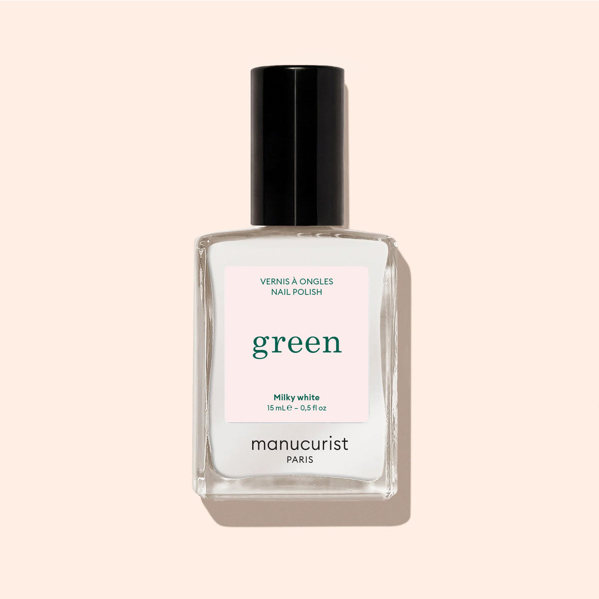 A clear glass bottle of Milky White nail polish by Manucurist with a black cap stands upright on a pale beige background.