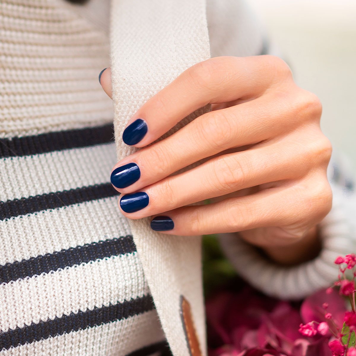 A hand with neatly manicured nails painted in Manucurist Navy Blue gel polish rests on a cream knit scarf. The person wears a white and navy striped sweater, with pink flowers partly visible in the bottom right corner.