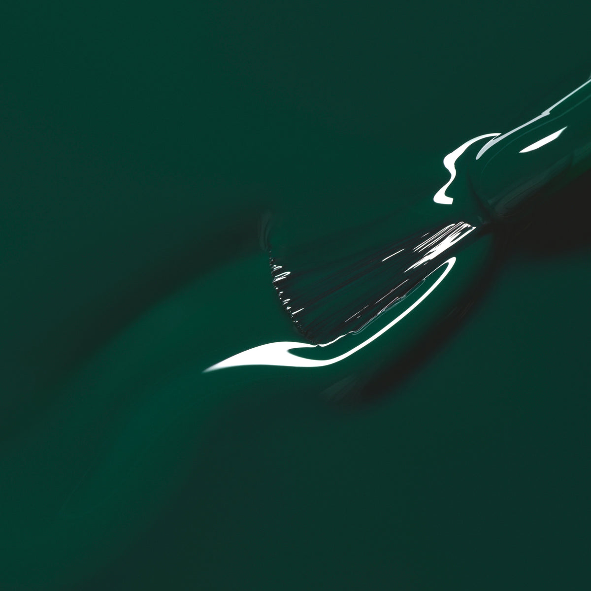 A close-up of a paintbrush spreading Manucurist’s Emerald nail polish, a glossy deep forest green shade, with light reflecting off the wet surface.