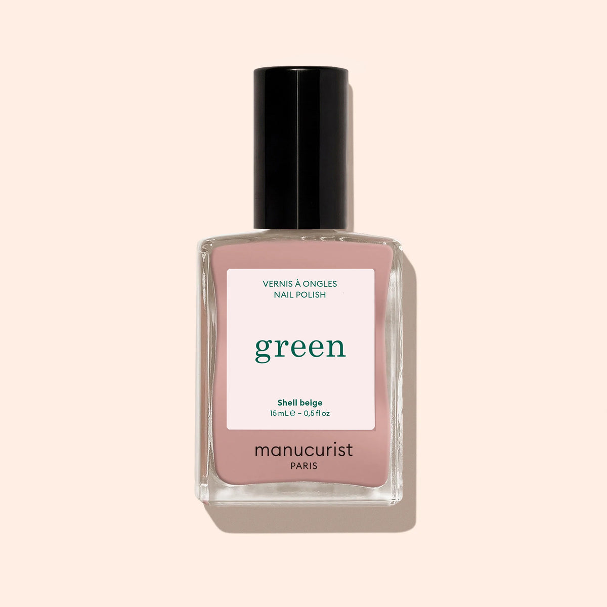 Un flacon de vernis à ongles Shell Beige de Manucurist, issu de la gamme Green™, avec un bouchon noir, un flacon en verre transparent et une étiquette beige-rose indiquant « green, quick dry intense color », sur fond beige clair.