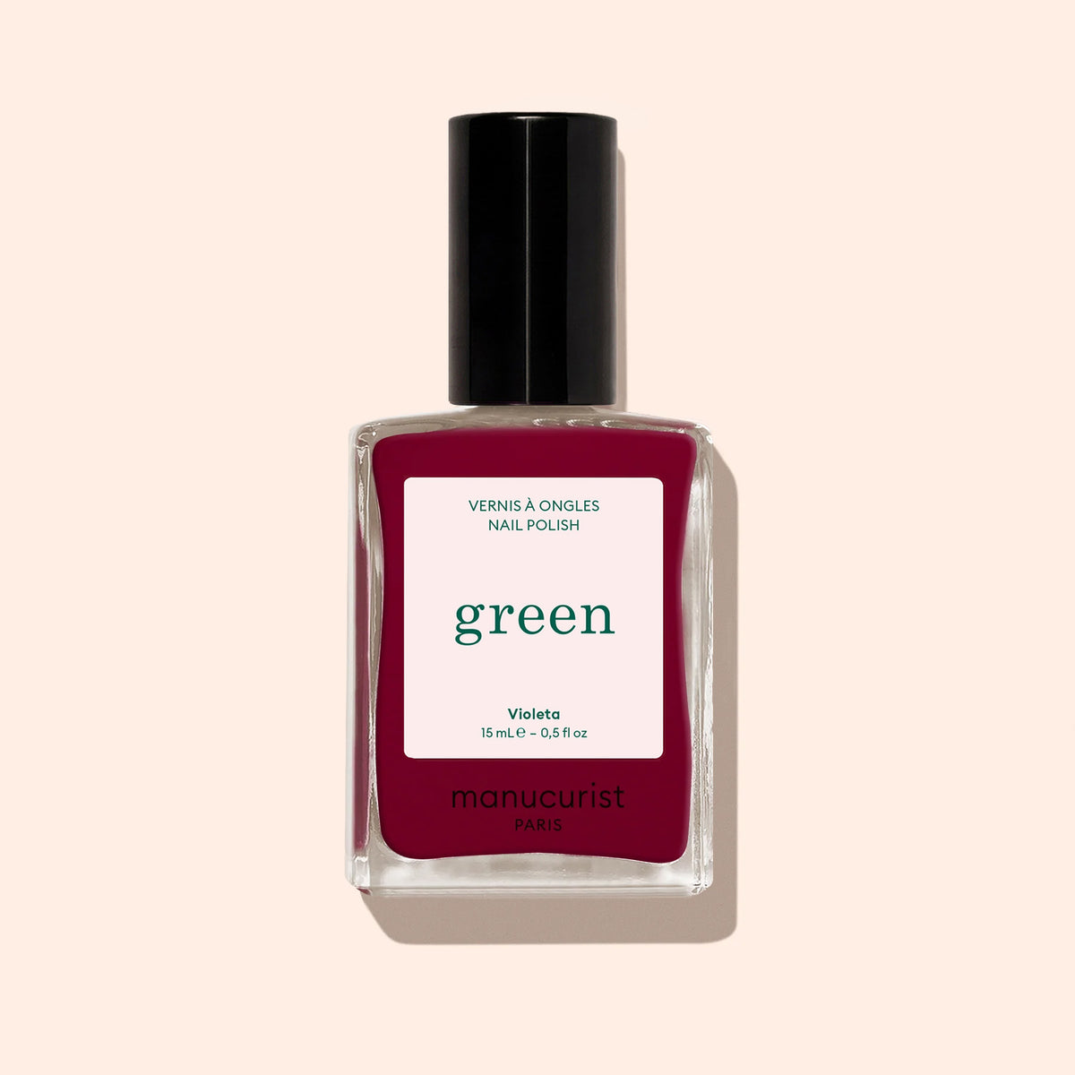 A clear bottle of Manucurist Violeta nail polish with a black cap, from the Green™ range, Quick Dry Intense Color. The deep burgundy purple shade stands out against a soft beige background.