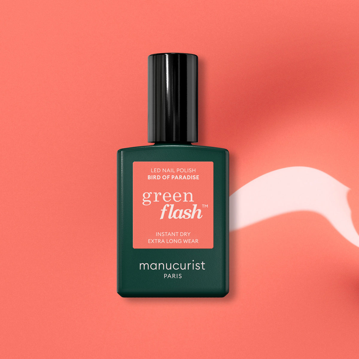 Un vernis gel orange corail éclatant, Bird of Paradise de Manucurist, est présenté dans un flacon vert sur un fond corail, avec à côté une traînée blanche façon peinture.