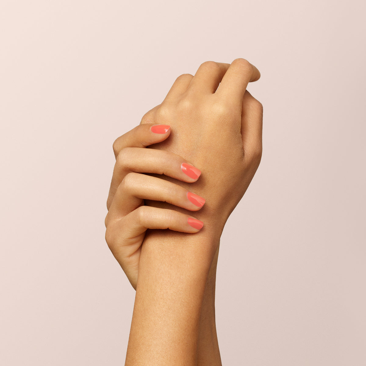 Gros plan sur deux mains croisées avec grâce sur fond beige clair, arborant des ongles impeccablement manucurés avec la teinte Bird of Paradise de Manucurist, un vernis gel orange corail brillant.