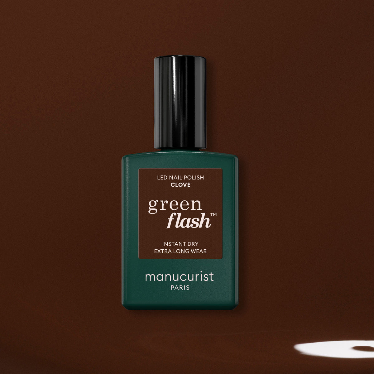A bottle of Manucurist Clove gel polish, a rich chocolate brown shade, is shown against a brown background with a small light spot near the lower right corner.