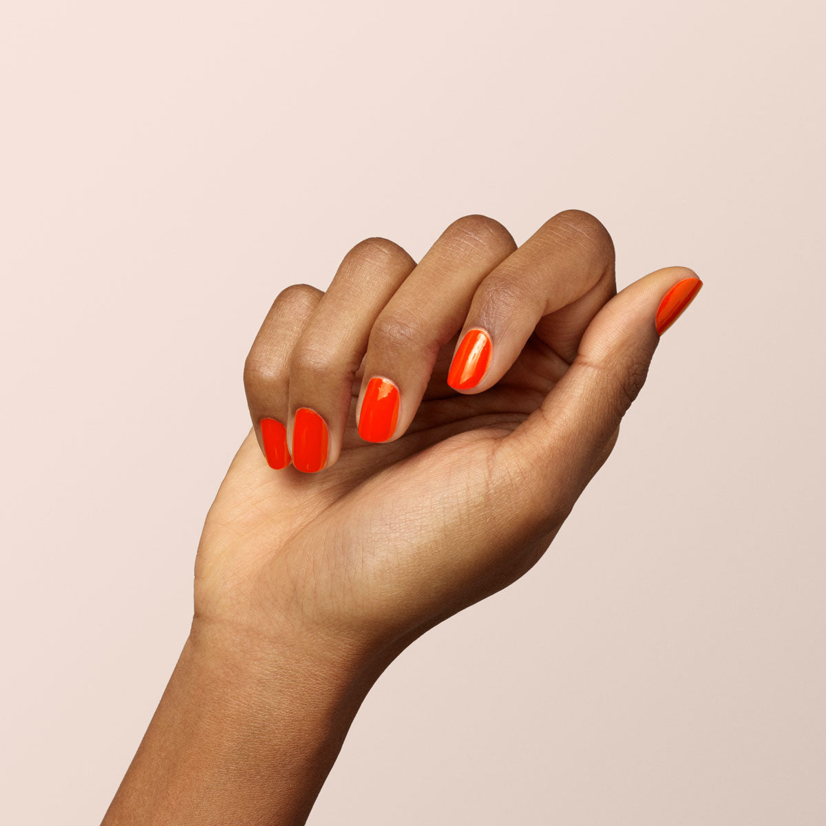 A hand with medium-brown skin is shown against a light beige background, nails neatly manicured and painted in Manucurist’s Coral Reef, a vibrant coral orange-red gel polish, with fingers gently curved to display the glossy finish.