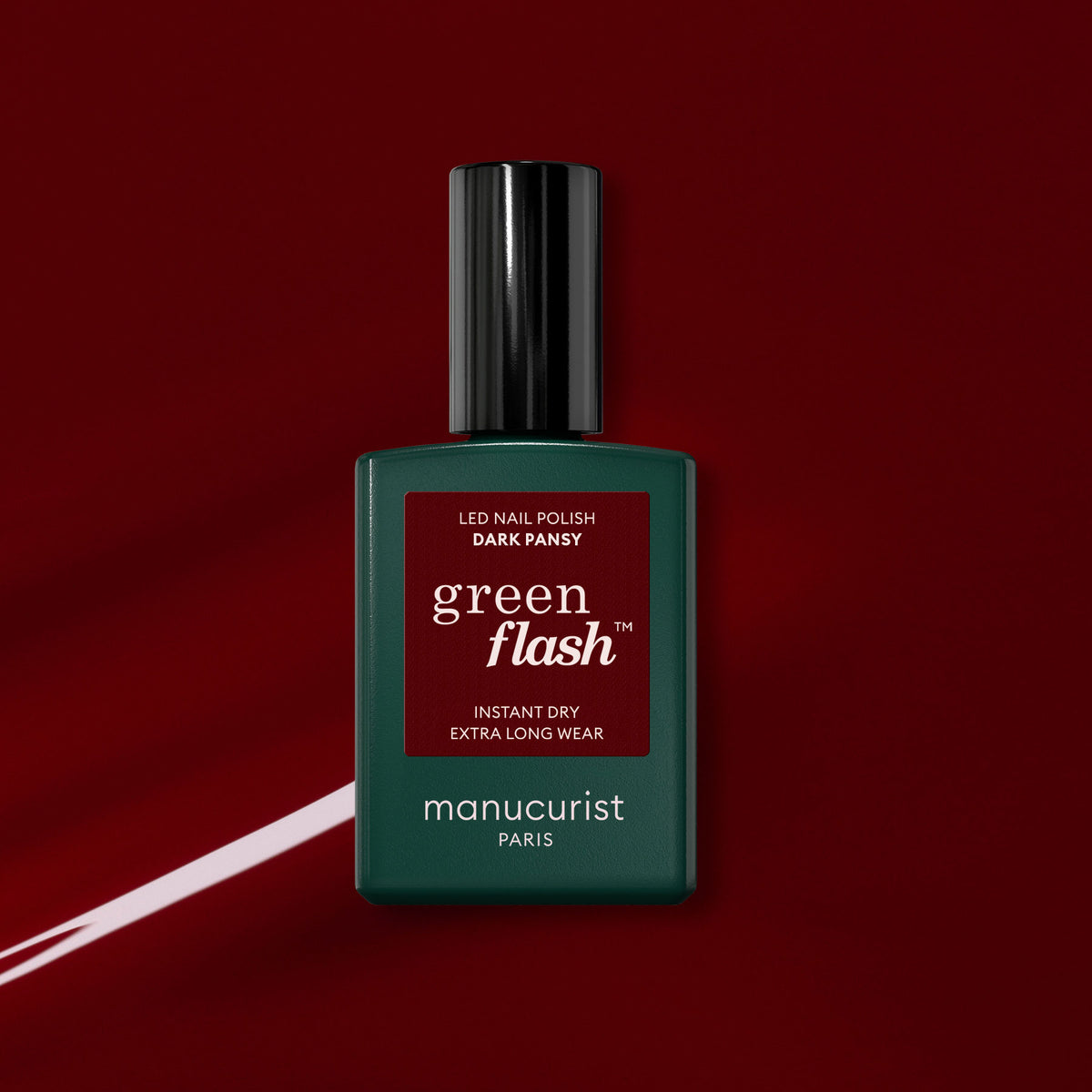 Un flacon de vernis gel Manucurist Dark Pansy repose sur un fond rouge profond et brillant, avec à côté une touche assortie de couleur pour ongles rouge velours.
