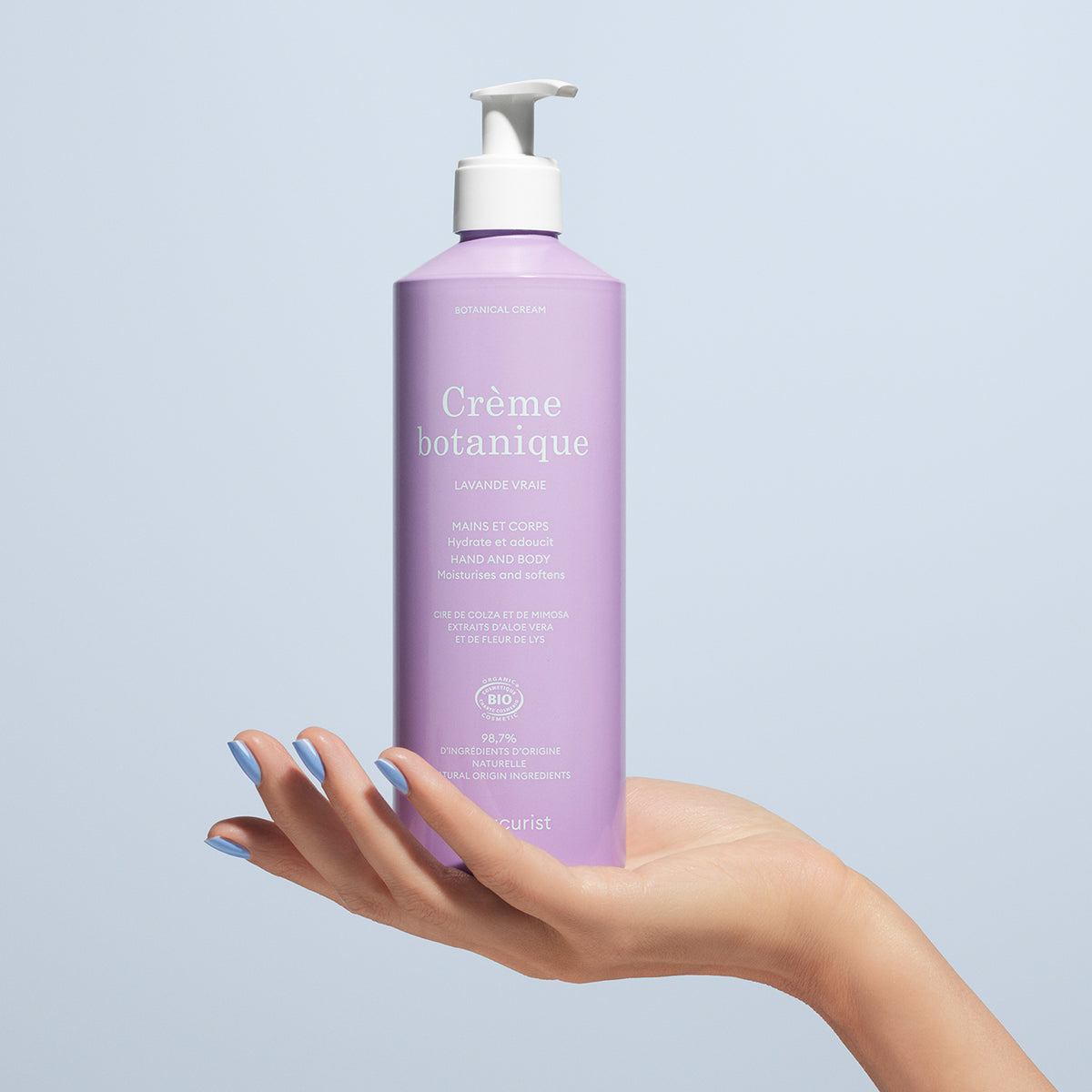Une main aux ongles vernis tient un flacon pompe lavande intitulé Soothing Lavender Hand Body Cream - Cosmos Organic by Manucurist, un soin hydratant bio pour les mains et le corps, sur fond bleu clair.
