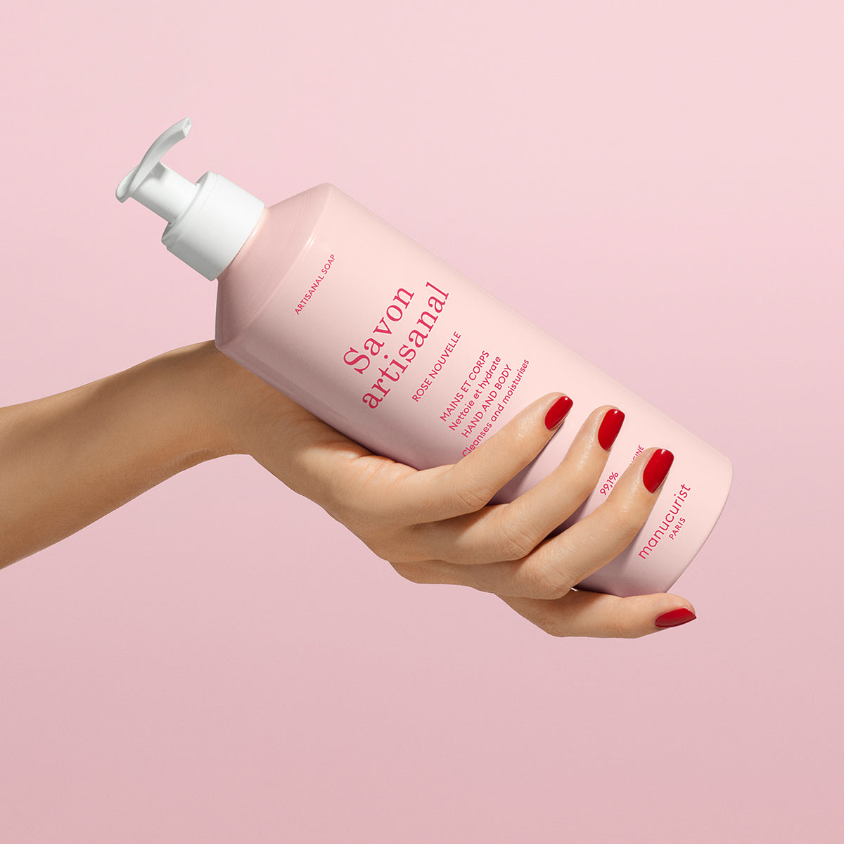 Une main aux ongles vernis rouges tient un flacon rose de gel lavant mains et corps Manucurist Organic Rose - Cosmos Organic avec une pompe blanche, sur un fond rose assorti.