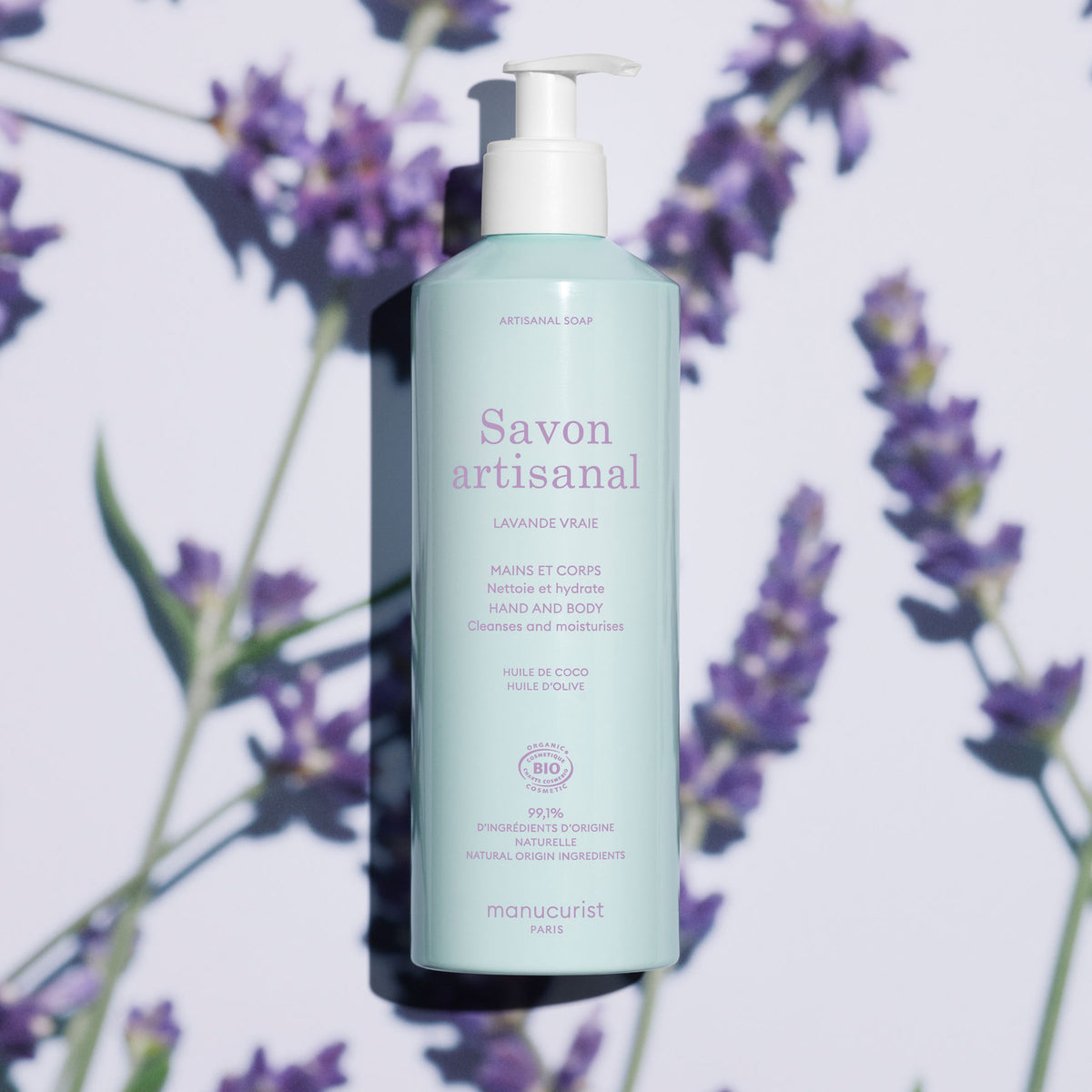 Un flacon bleu clair de Gel Lavant Mains et Corps Lavande Bio Manucurist - Cosmos Organic avec pompe repose sur une surface avec des fleurs de lavande et des ombres douces. L&