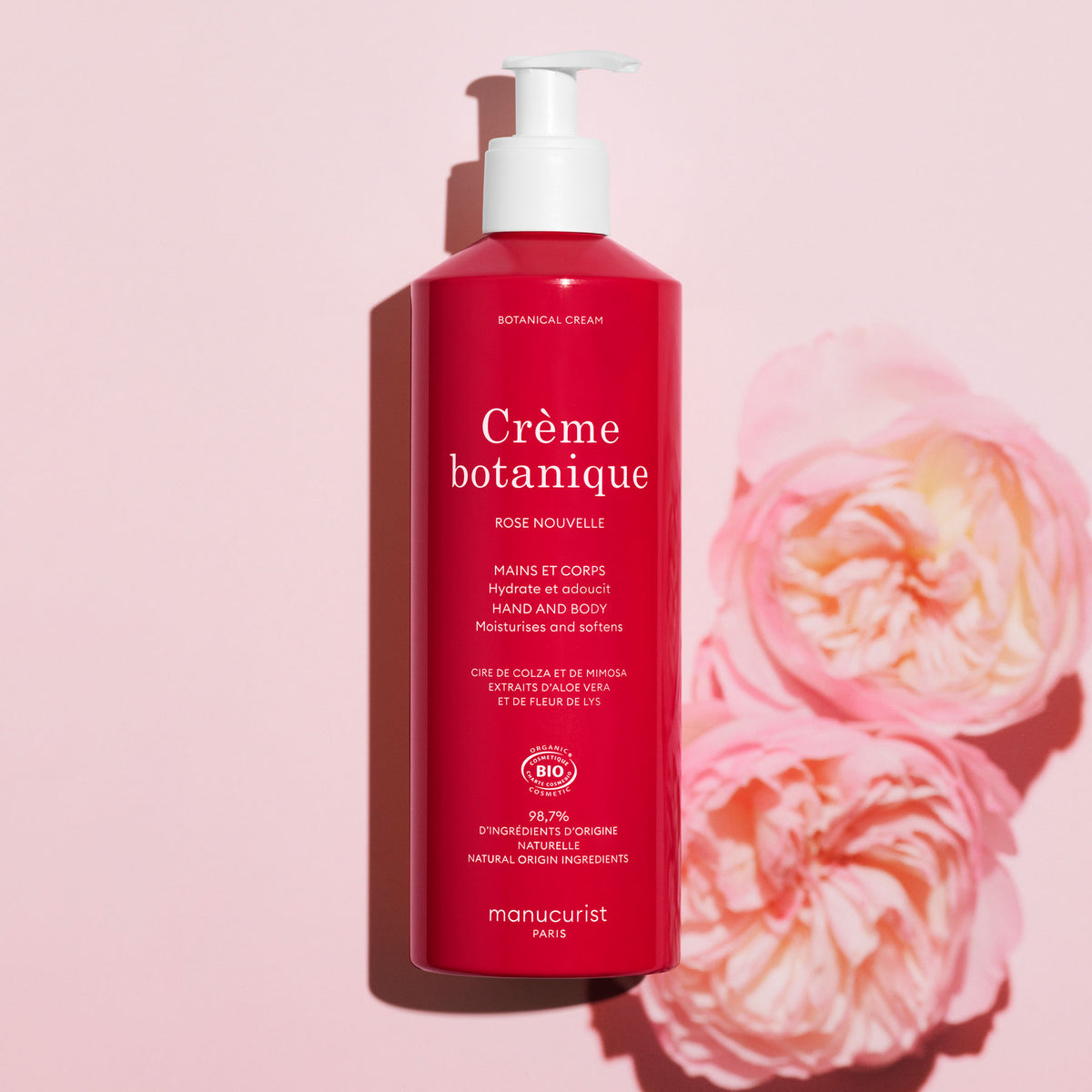 Un flacon rose de crème pour les mains et le corps Soothing Rose de Manucurist - Cosmos Organic se tient debout sur un fond rose clair, à côté de deux roses roses. La pompe blanche et le texte mettent en avant sa promesse d’hydrater profondément votre peau.