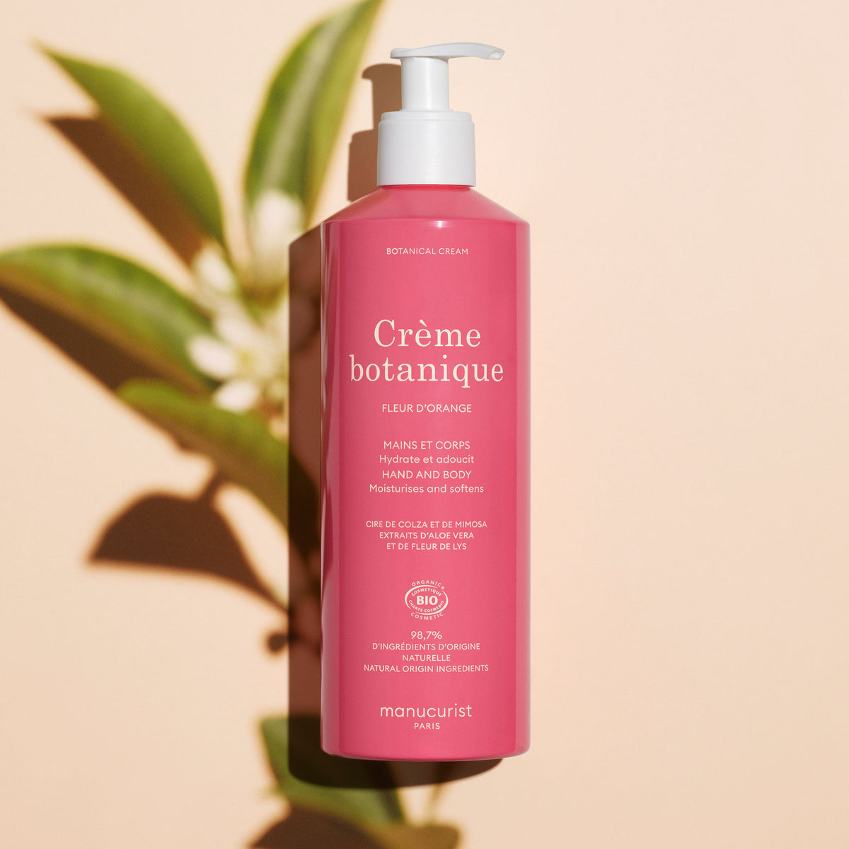 Un flacon rose de Soothing Orange Blossom Hand Body Cream – Cosmos Organic Manucurist avec pompe est posé sur une surface claire, projetant une ombre de feuillage vert. L&