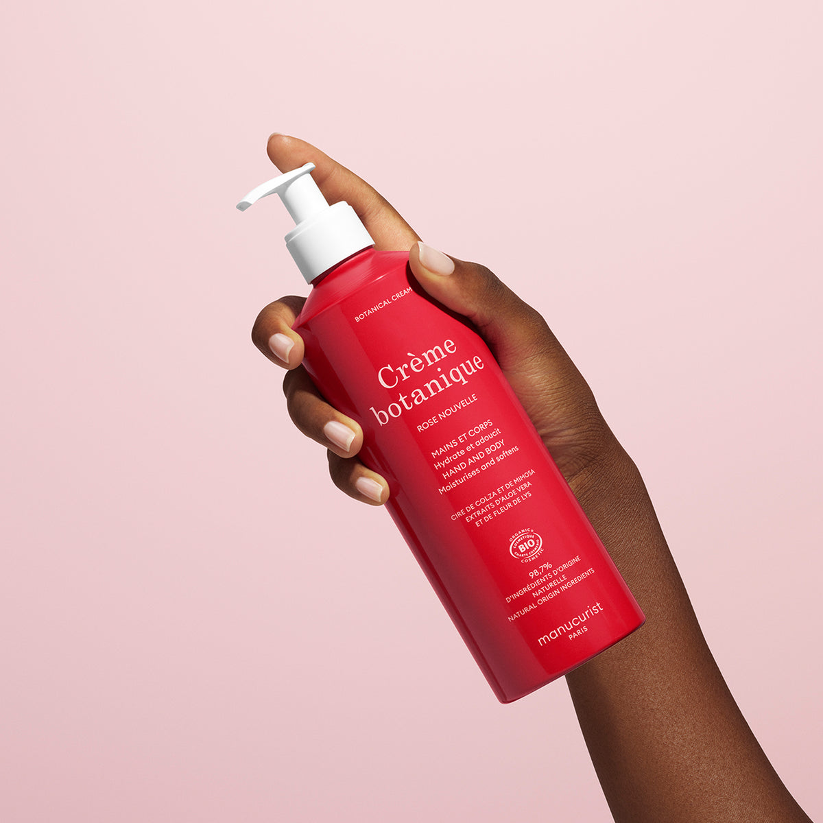 Une main tient un flacon rouge de Crème Mains & Corps Rose Apaisante - Cosmos Organic Manucurist doté d&