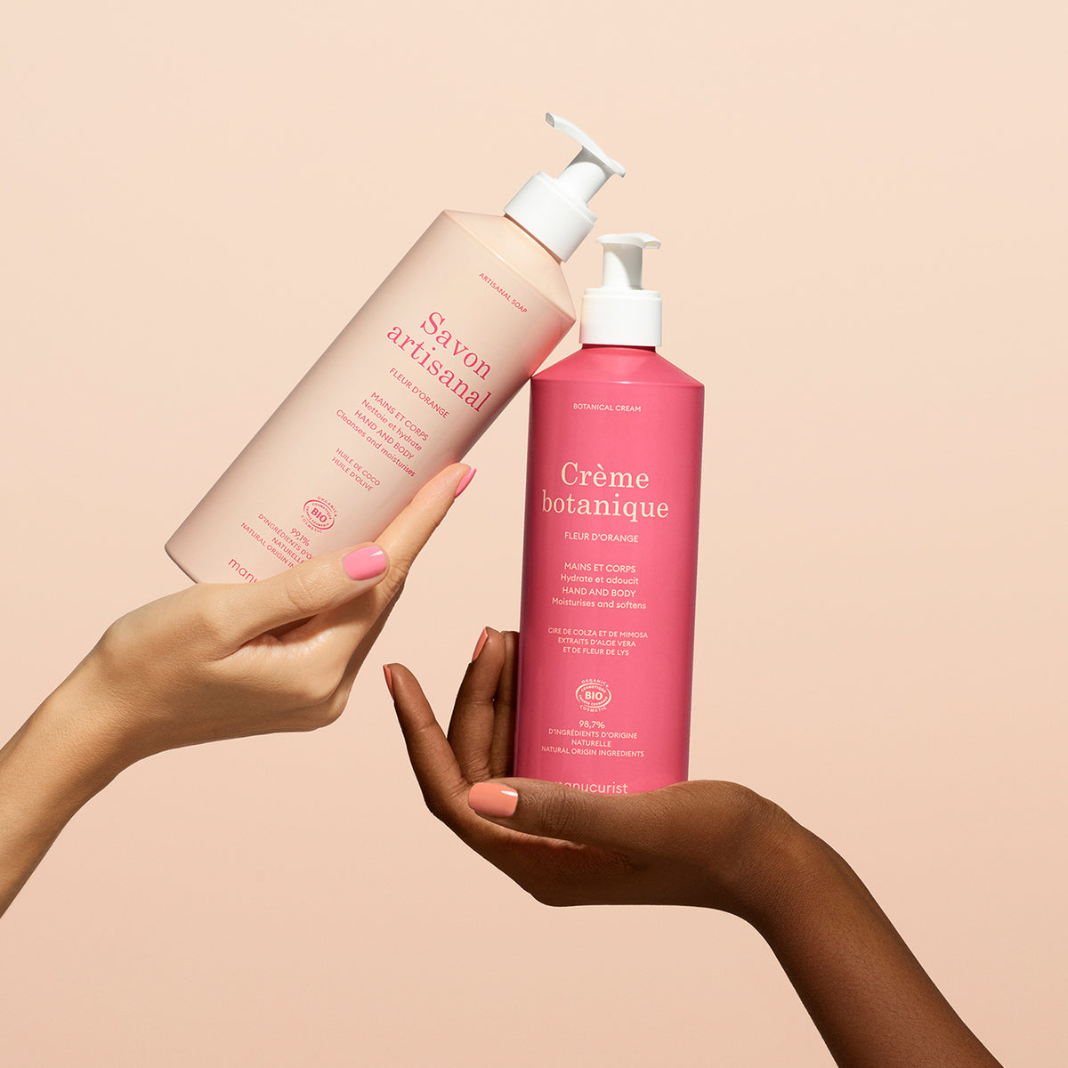Two hands hold up Manucurist’s Orange Blossom Botanical Cream Artisan Soap Duo - Cosmos Organic against a light peach background: the left holds the artisan soap, while the right displays the bright pink botanical cream.