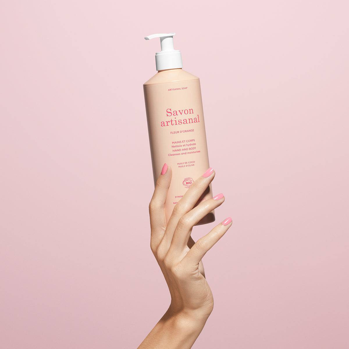 Une main manucurée tient un flacon rose de Manucurist Gel lavant mains et corps à la fleur d’oranger bio – Cosmos Organic, sur fond rose pâle.