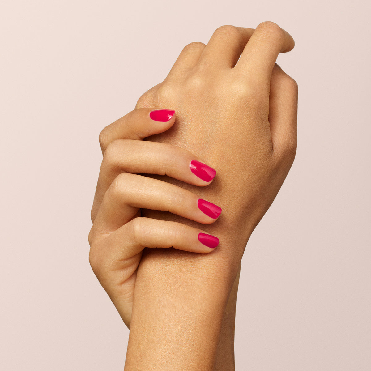 A close-up of two hands with neatly manicured nails painted in Manucurist Péonie, a vibrant raspberry pink gel polish, set against a soft, light beige background.