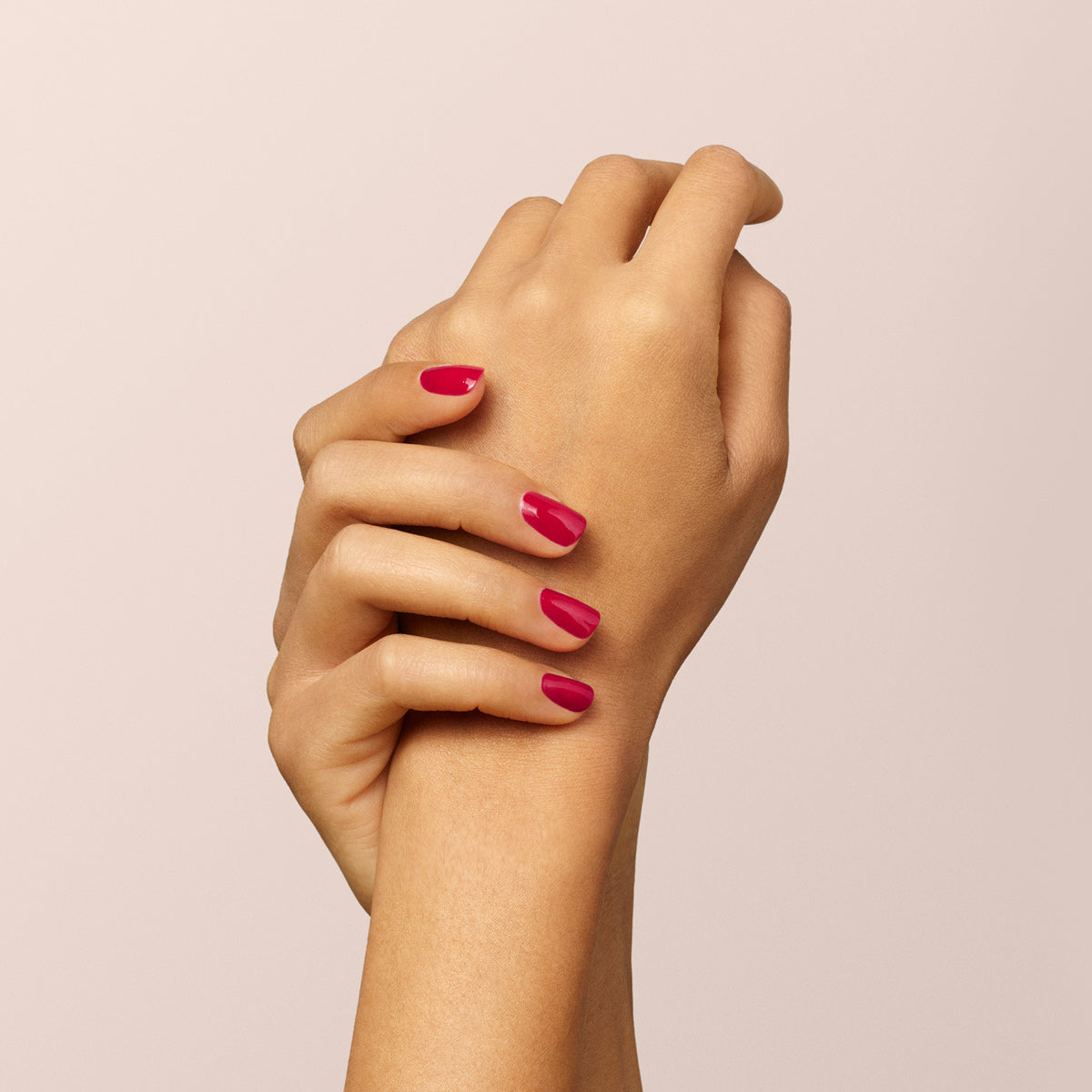 A close-up of two hands clasped together, showcasing neatly manicured nails painted in Manucurist’s vivid Pomegranate shade against a pale background.