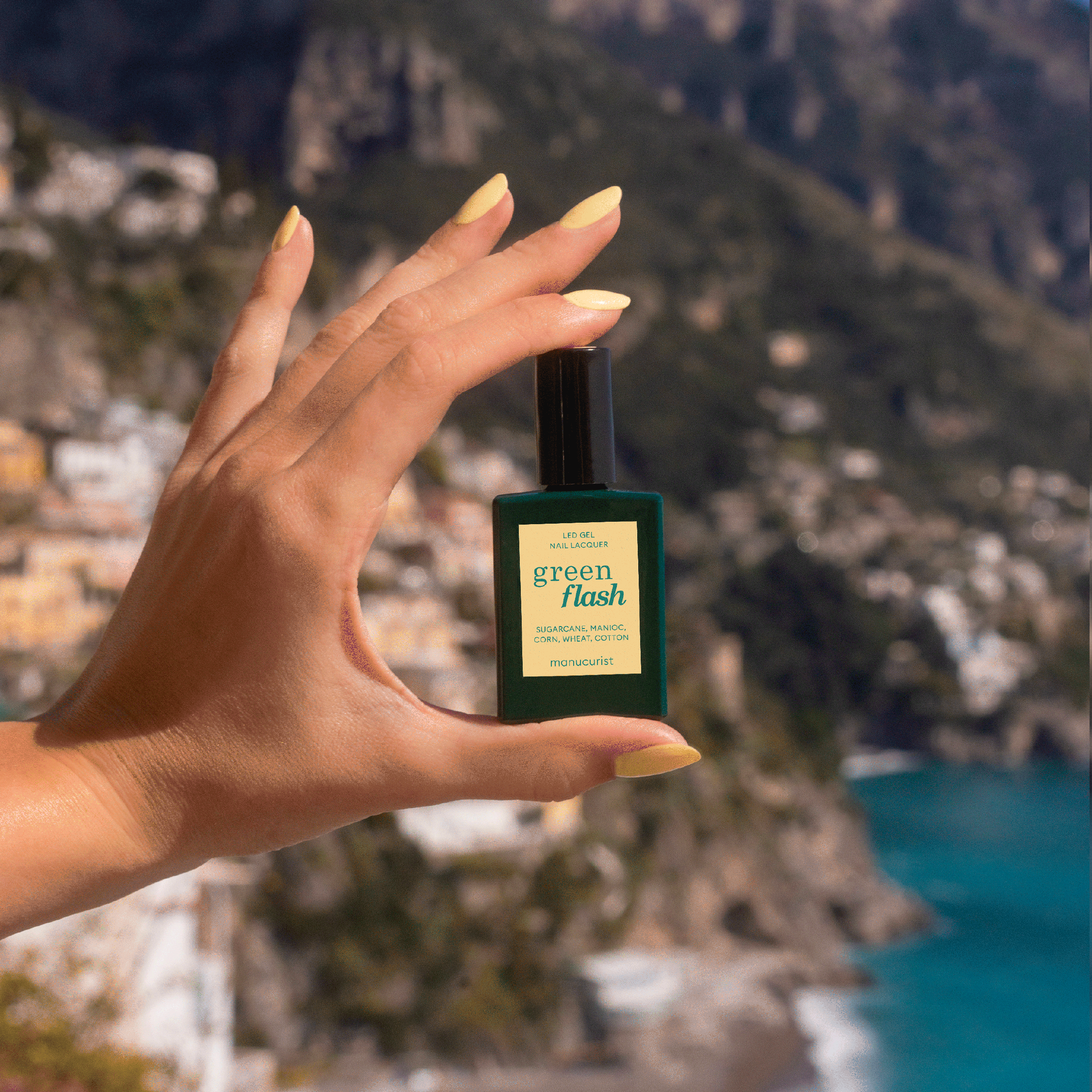 A hand with pastel yellow gel polish in shade Mimosa by Manucurist holds a bottle of Green Flash gel polish against a blurred coastal town backdrop with cliffs, buildings, and the sea.