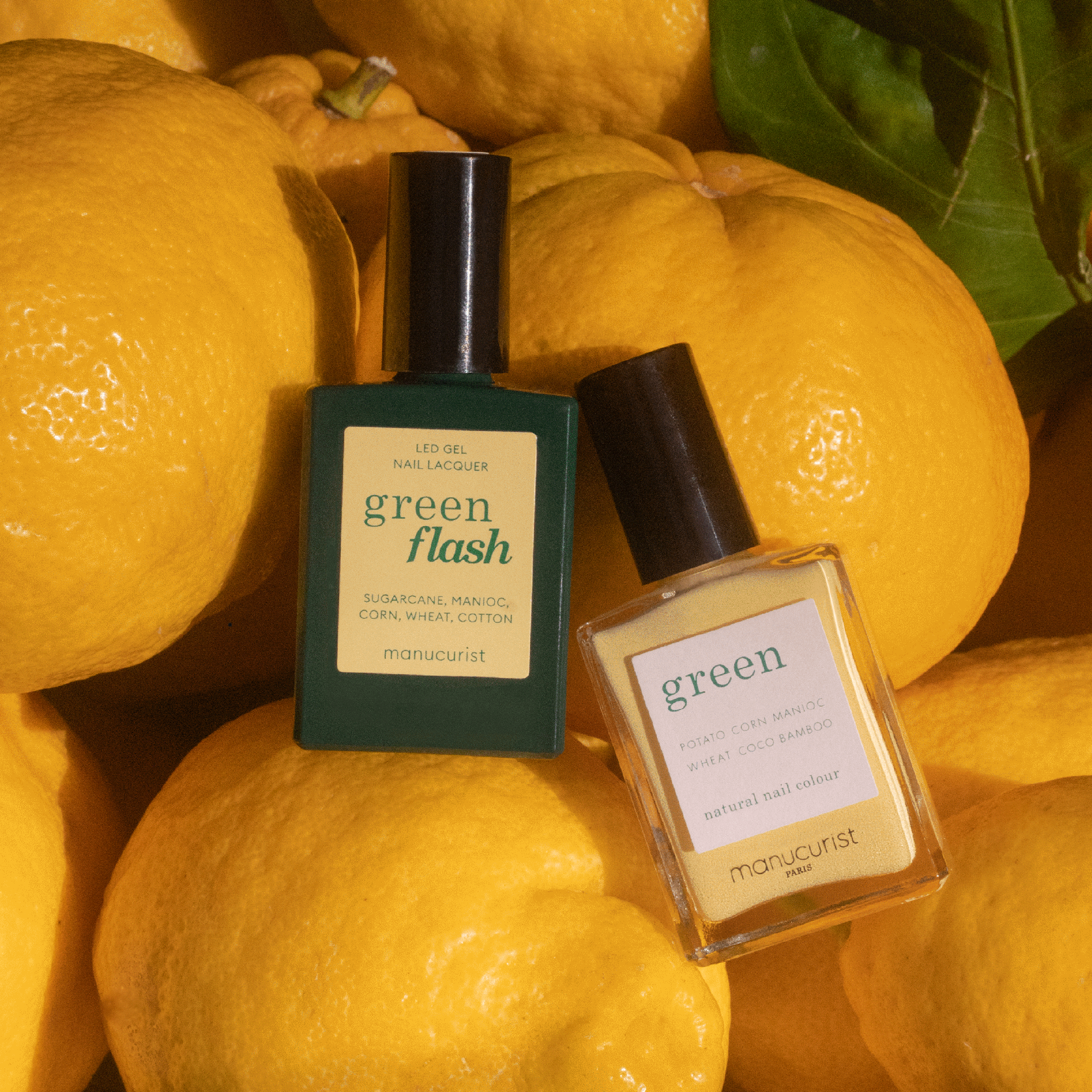 Two bottles of Manucurist nail polish in the shade Mimosa are displayed on a pile of bright yellow lemons with green leaves.