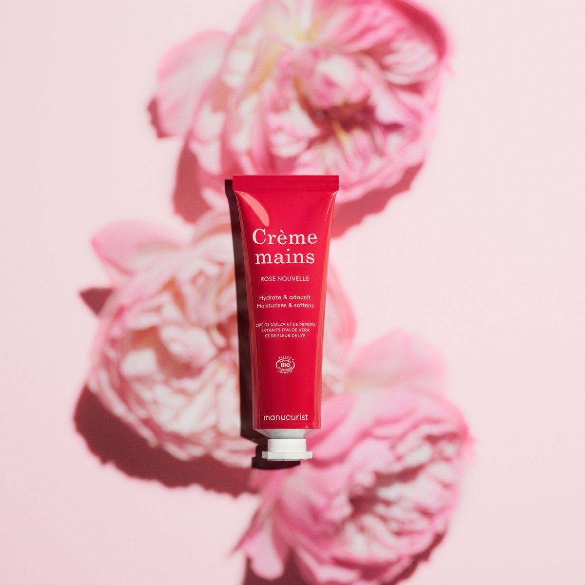 Un tube rouge de Crème Mains Rose Quotidienne - Cosmos Organic par Manucurist est centré sur un fond rose avec trois fleurs roses floutées projetant des ombres. Le produit offre une délicieuse senteur de rose.