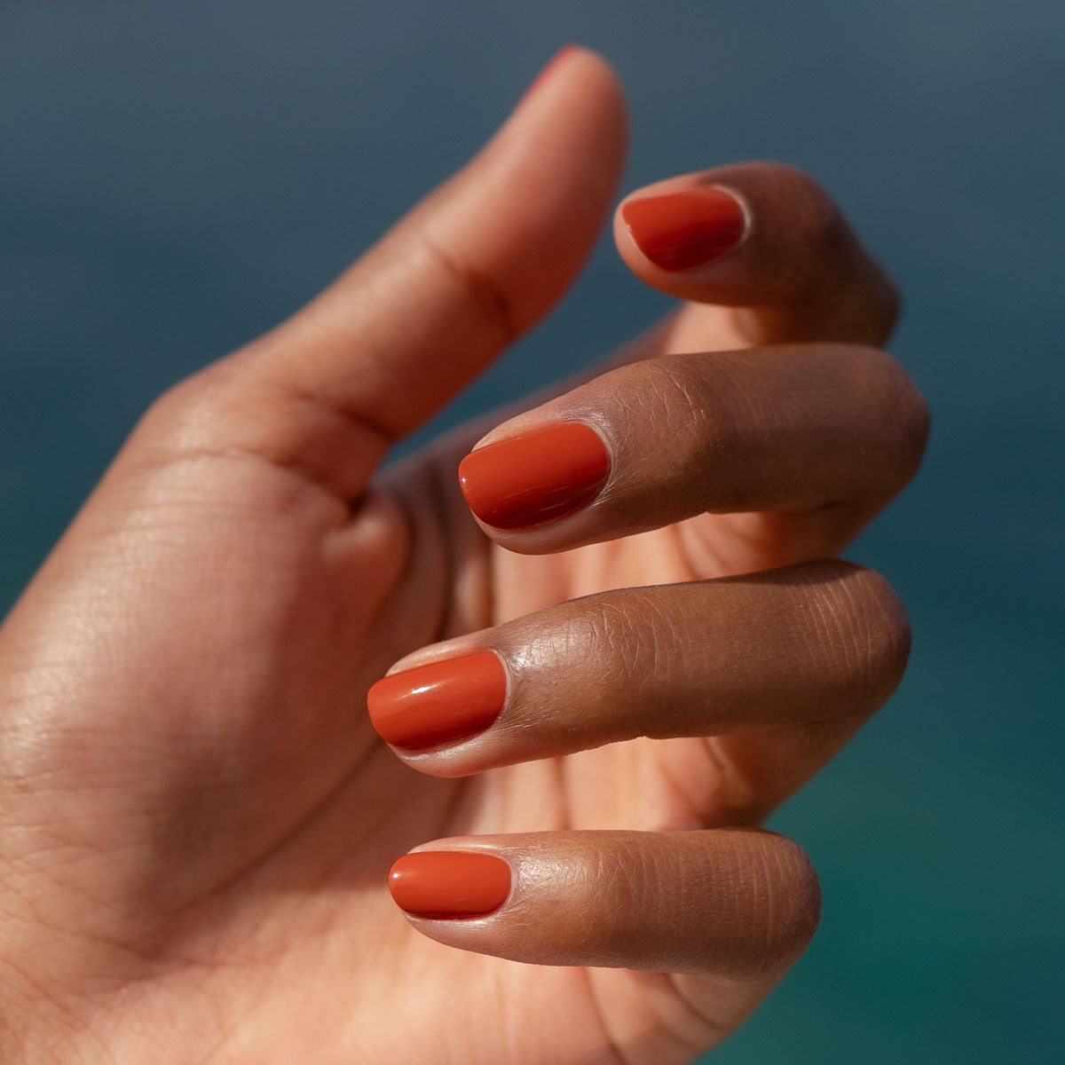 A close-up of a medium brown hand with neatly manicured nails painted in Manucurist’s Green Flash gel polish in the Terracotta shade, a glossy burnt orange, set against a blurred blue background.
