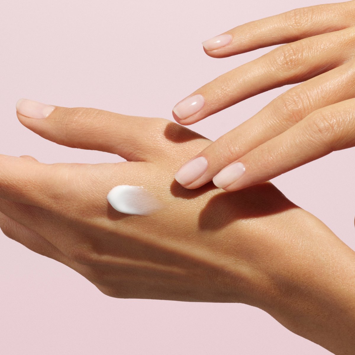 Gros plan d’une main aux ongles manucurés appliquant une petite quantité de crème pour les mains Manucurist Everyday Rose - Cosmos Organic sur le dos de l’autre main, sur fond rose poudré.