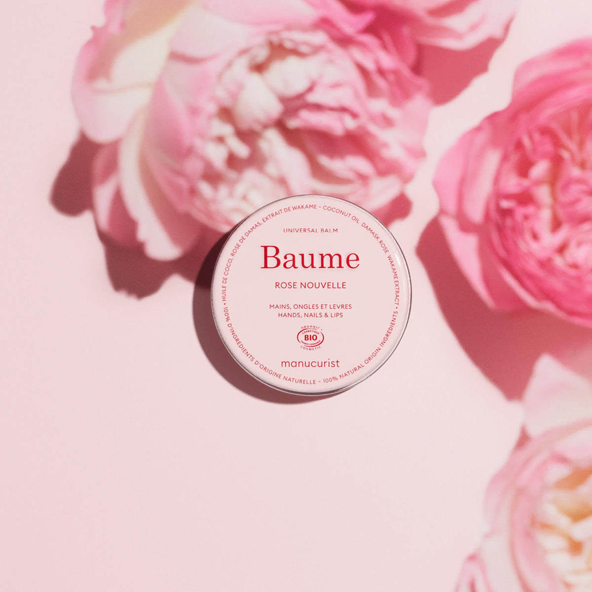 Un pot rond de Baume Nourrissant Naturel Manucurist - Cosmos Organic repose sur une surface rose clair, entouré de douces fleurs roses, créant un univers délicat et féminin.