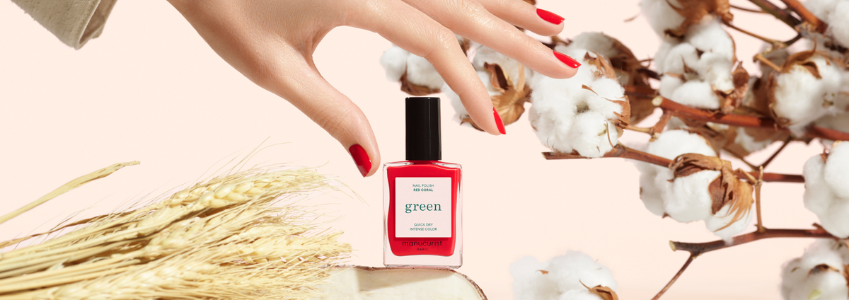 A hand with red nail polish reaches toward a red nail polish bottle next to cotton branches, wheat, and a blue egg labeled M7XQ in a straw nest on a beige background.