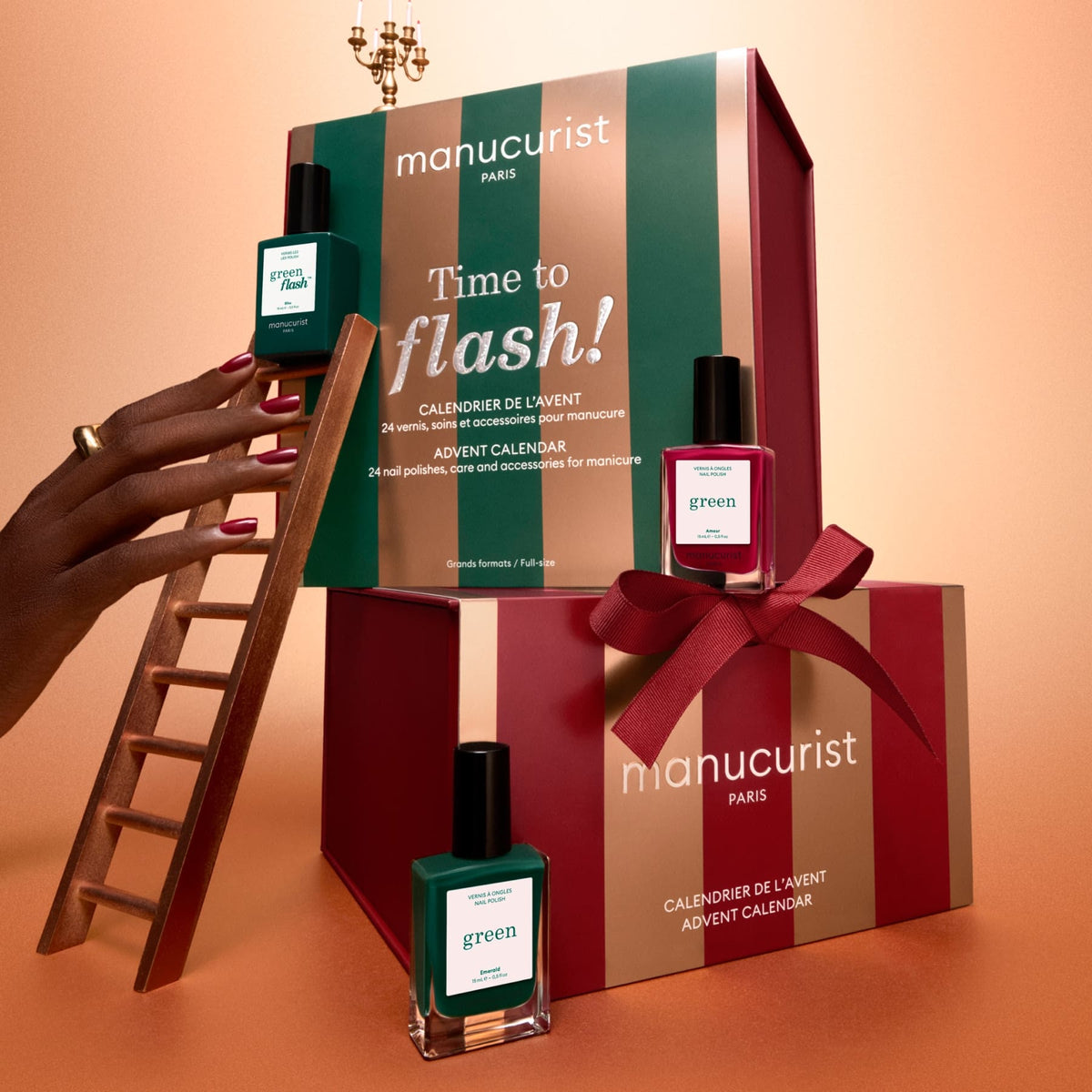 A hand with dark red nails holds a small ladder next to boxed Manucurist advent calendars and three bottles of green nail polish on a peach-colored background.