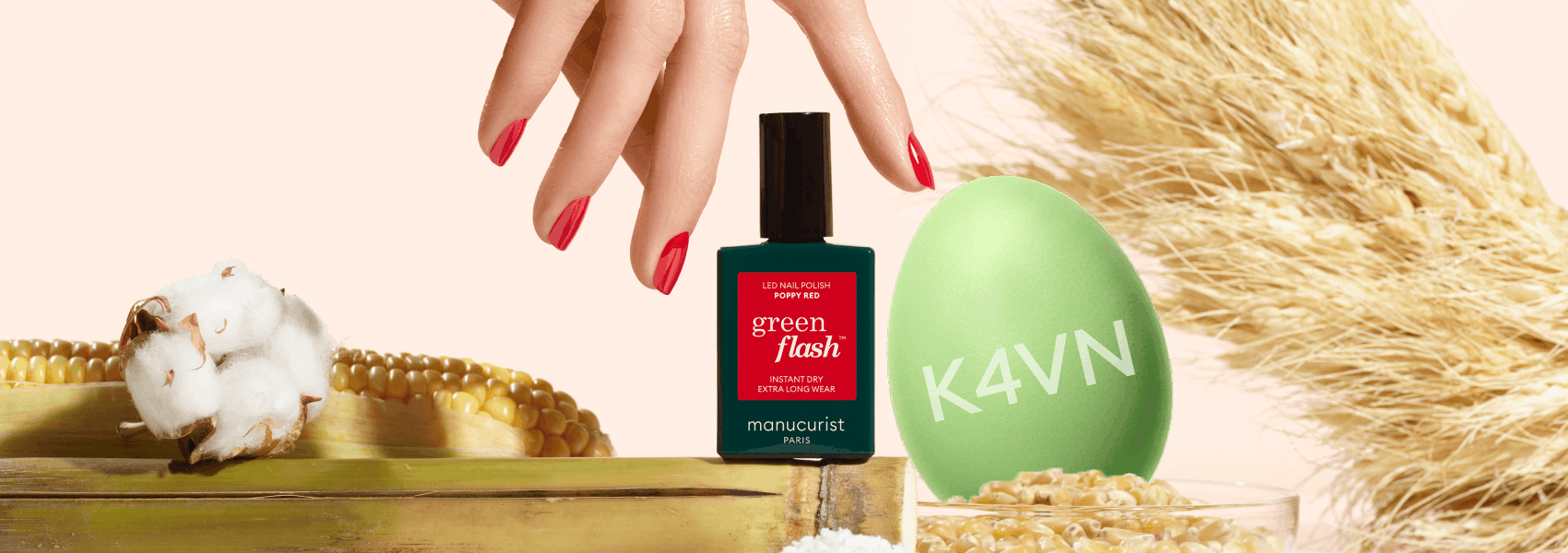 A hand with red-painted nails hovers above a green nail polish bottle labeled green flash. Surrounding it are a green K4VN egg, dried wheat, cotton, corn, and grains on a light background.