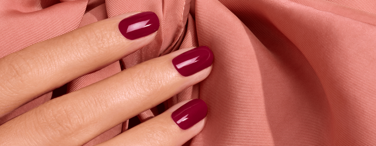Hand with fingernails painted a deep, glossy berry red resting on draped peach-colored fabric.